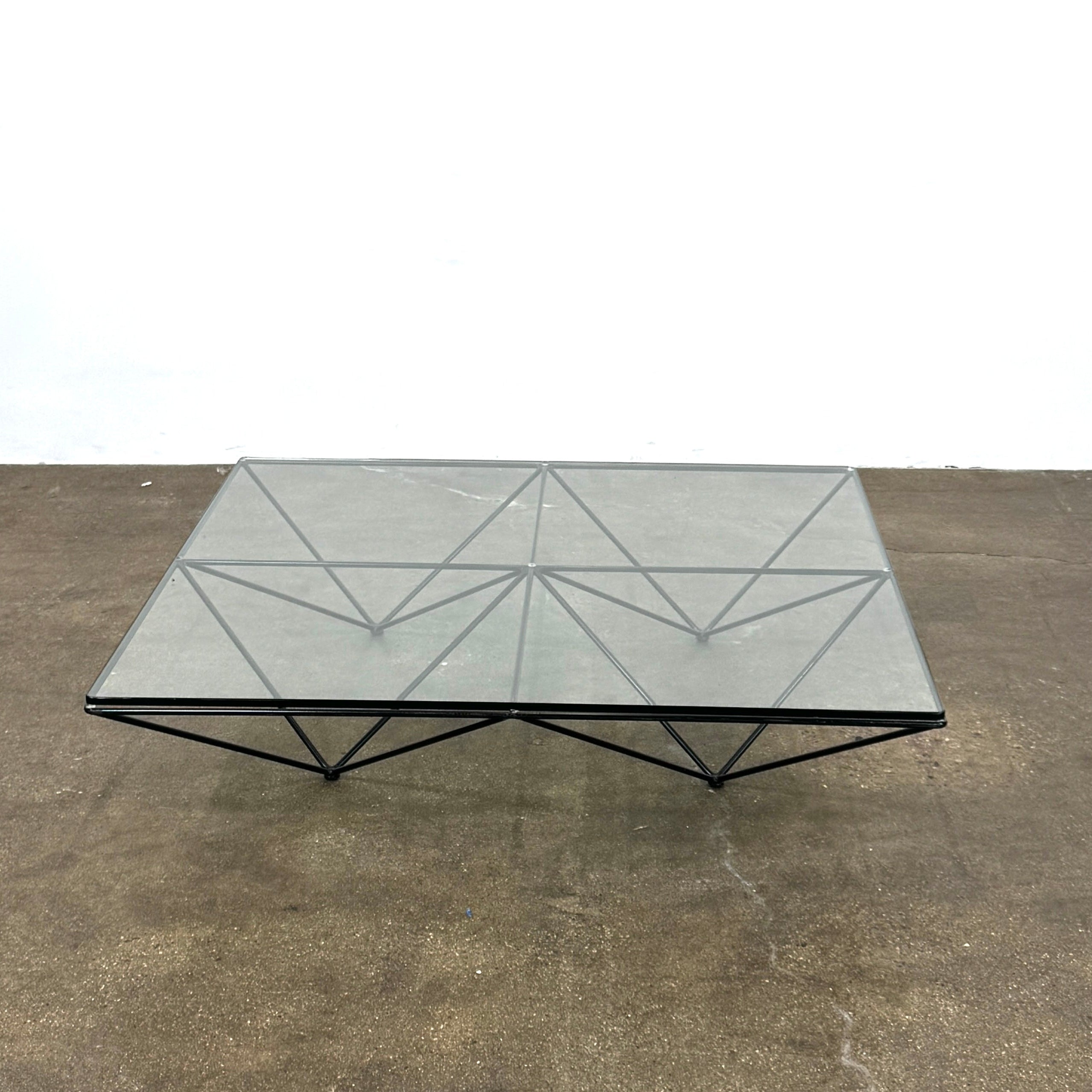 A B&B Italia Alanda Coffee Table with a glass top and metal frame sits on a polished concrete floor, with a magazine placed on its corner.