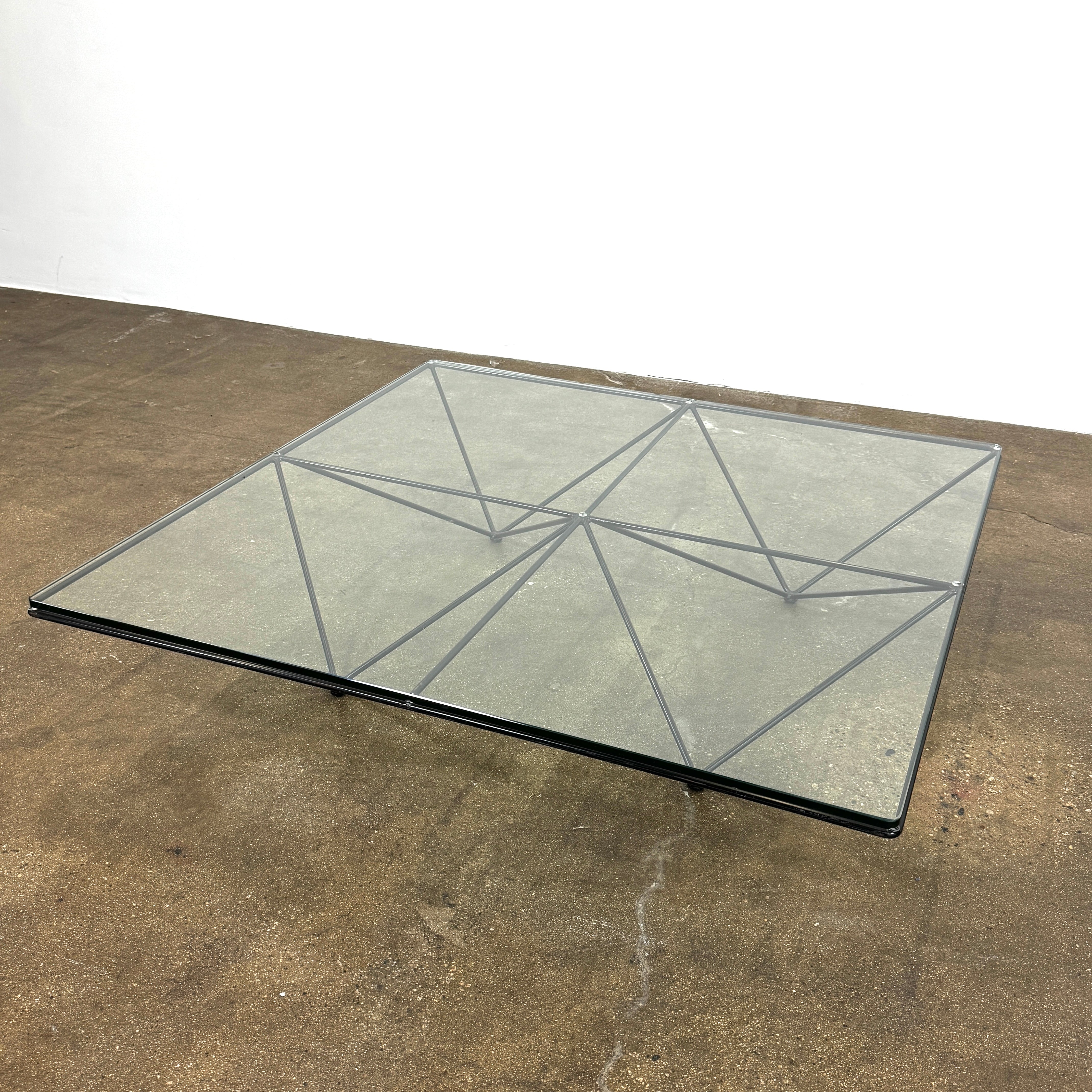 A B&B Italia Alanda Coffee Table with a glass top and metal frame sits on a polished concrete floor, with a magazine placed on its corner.