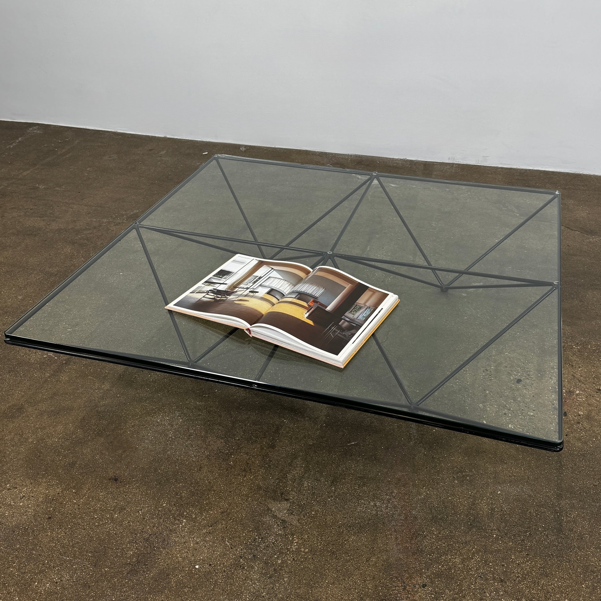 A B&B Italia Alanda Coffee Table with a glass top and metal frame sits on a polished concrete floor, with a magazine placed on its corner.