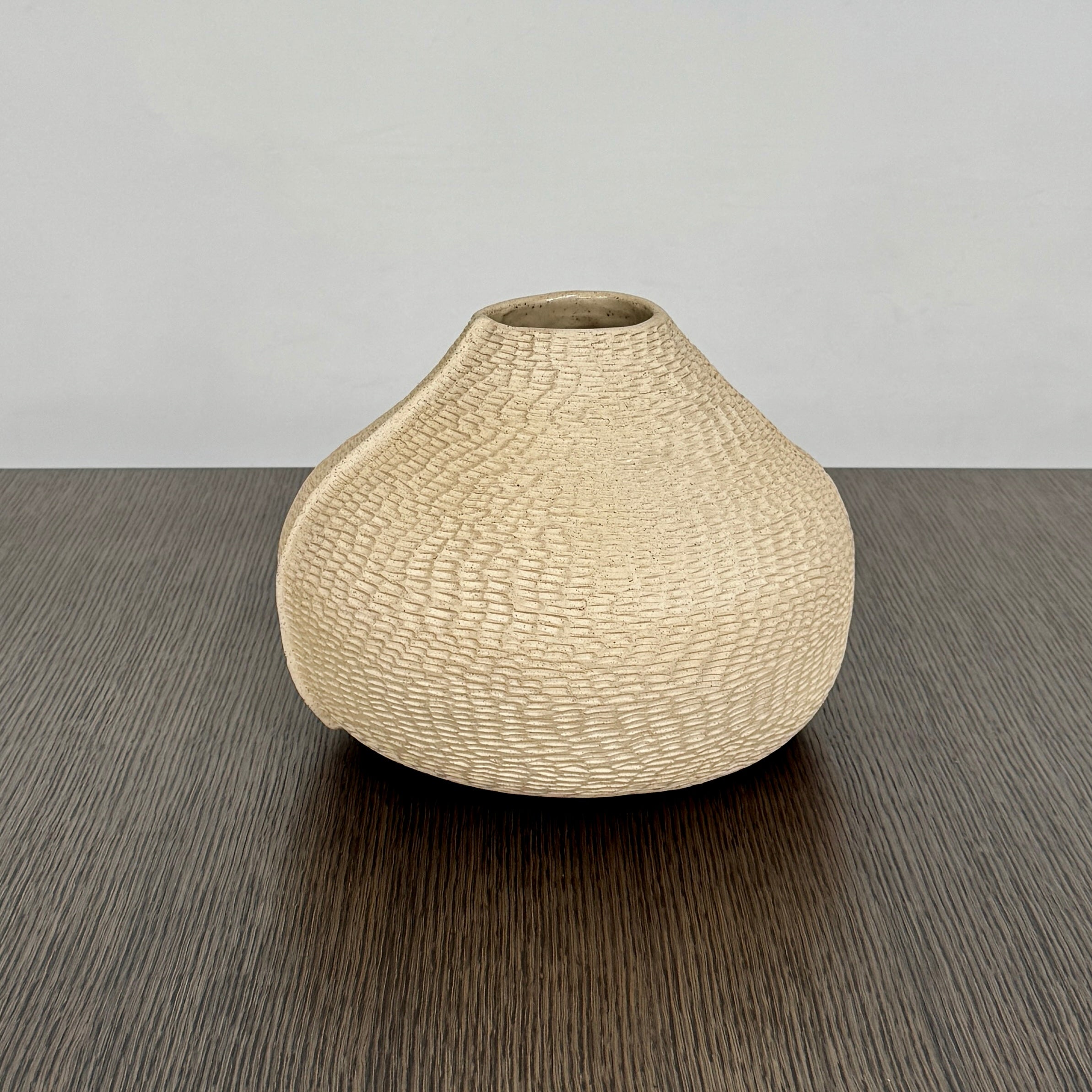 The Sofi Ceramic Pleated Light Clay Vase by Artist, featuring a textured, sculptural form and small opening, sits atop a dark wood-grain surface.