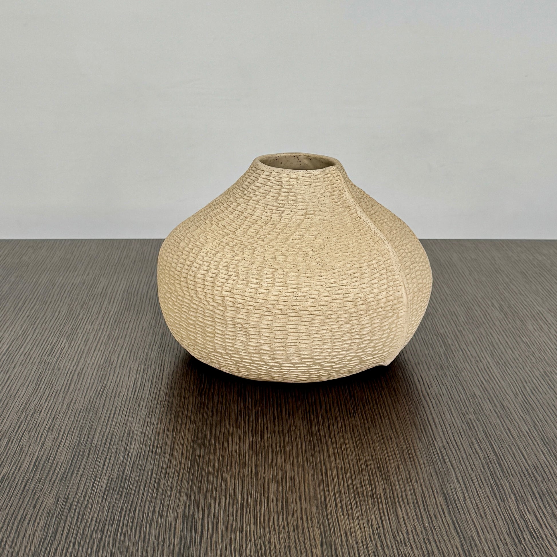 The Sofi Ceramic Pleated Light Clay Vase by Artist, featuring a textured, sculptural form and small opening, sits atop a dark wood-grain surface.