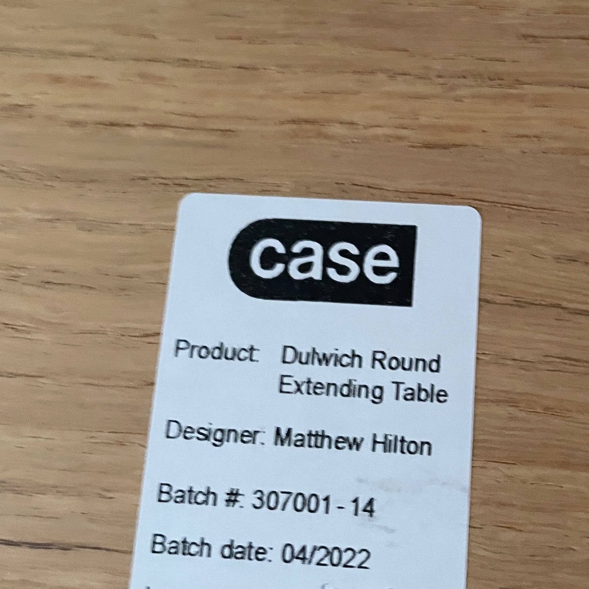 A Case Dulwich Extension Table, round and wooden with four legs, stands on a concrete floor against a plain white wall.