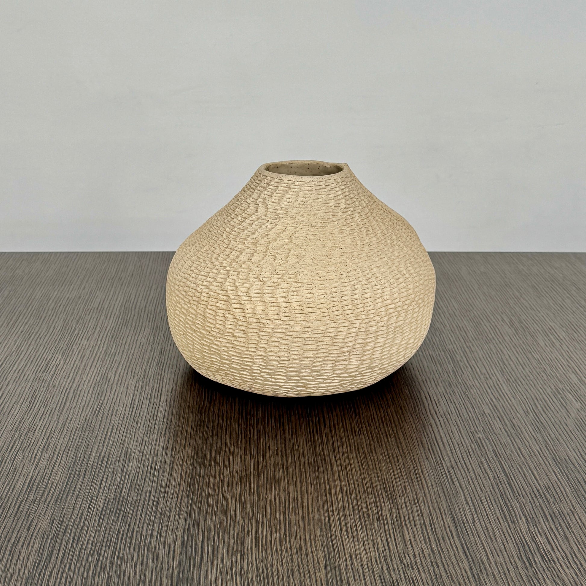 The Sofi Ceramic Pleated Light Clay Vase by Artist, featuring a textured, sculptural form and small opening, sits atop a dark wood-grain surface.