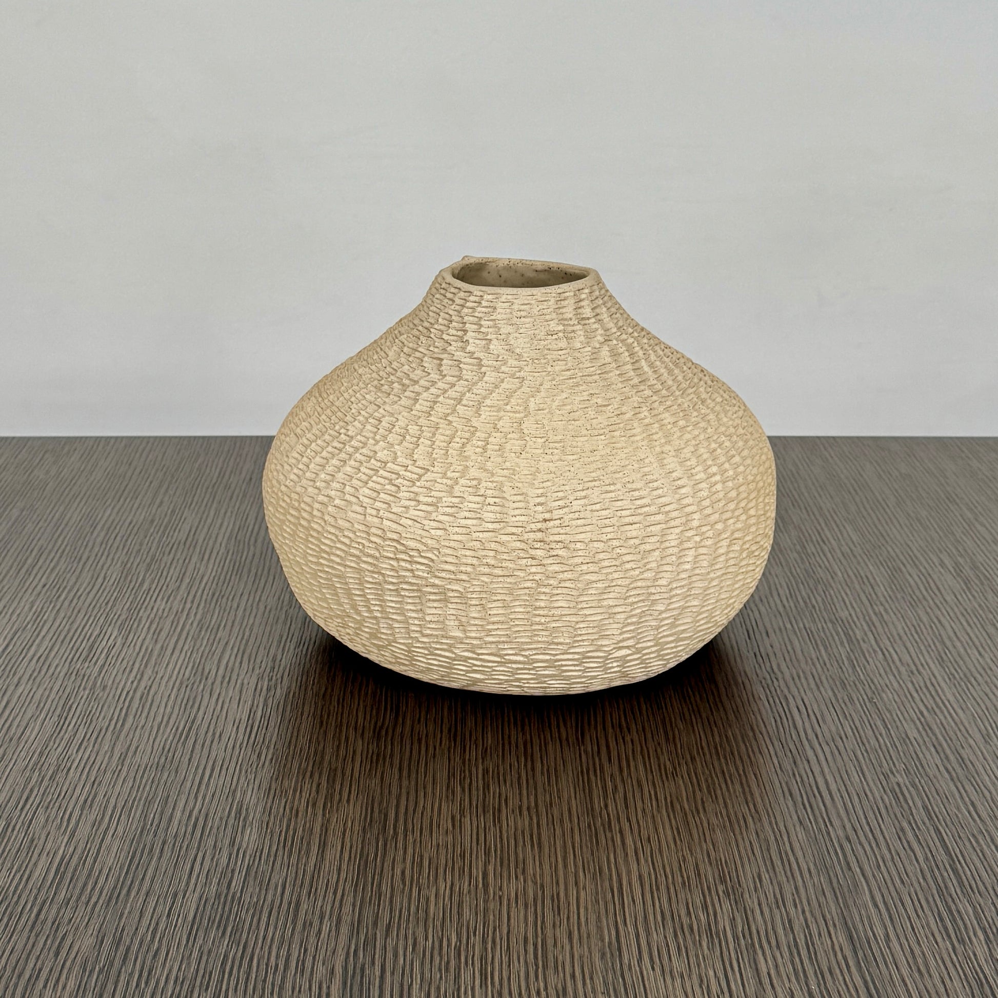 The Sofi Ceramic Pleated Light Clay Vase by Artist, featuring a textured, sculptural form and small opening, sits atop a dark wood-grain surface.