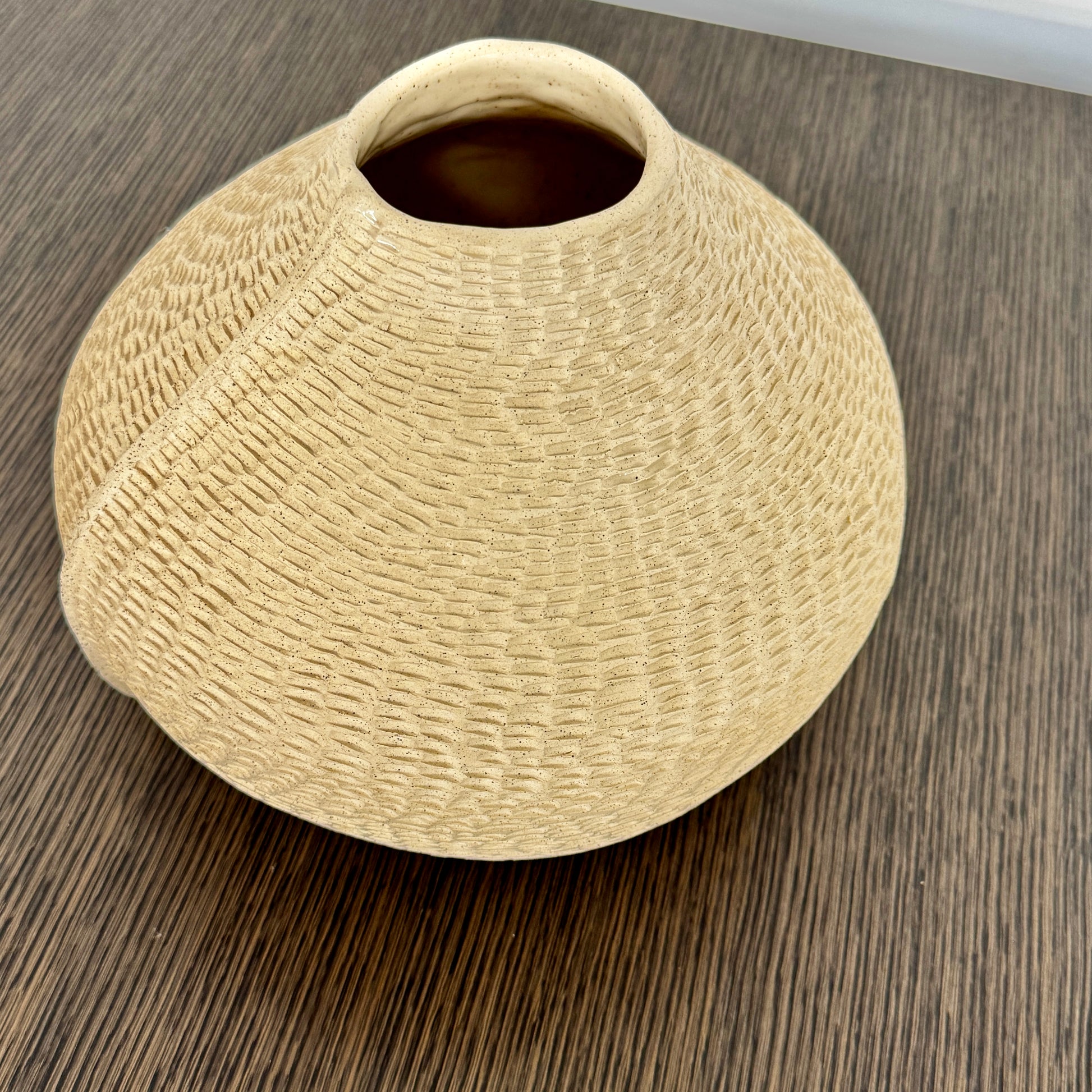 The Sofi Ceramic Pleated Light Clay Vase by Artist, featuring a textured, sculptural form and small opening, sits atop a dark wood-grain surface.