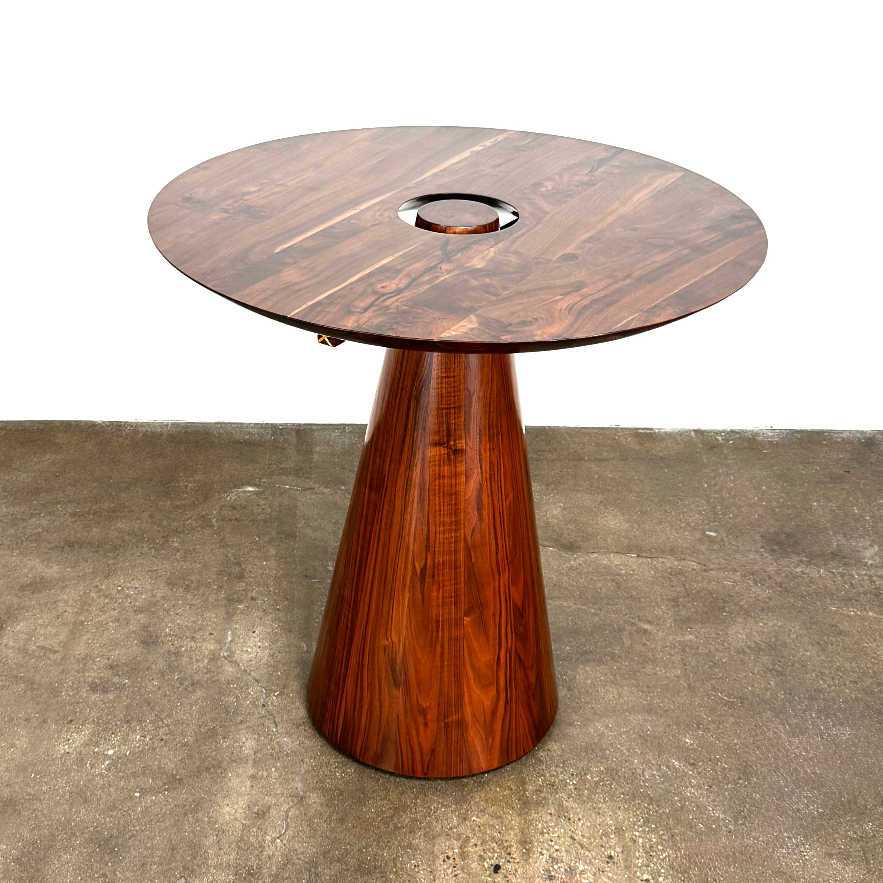 The Mike Fair Claro Walnut Bar Height Table by Artist features a round claro walnut top with a central circular cutout, a conical base, and a small gold cross detail on the side, shown on a concrete floor.