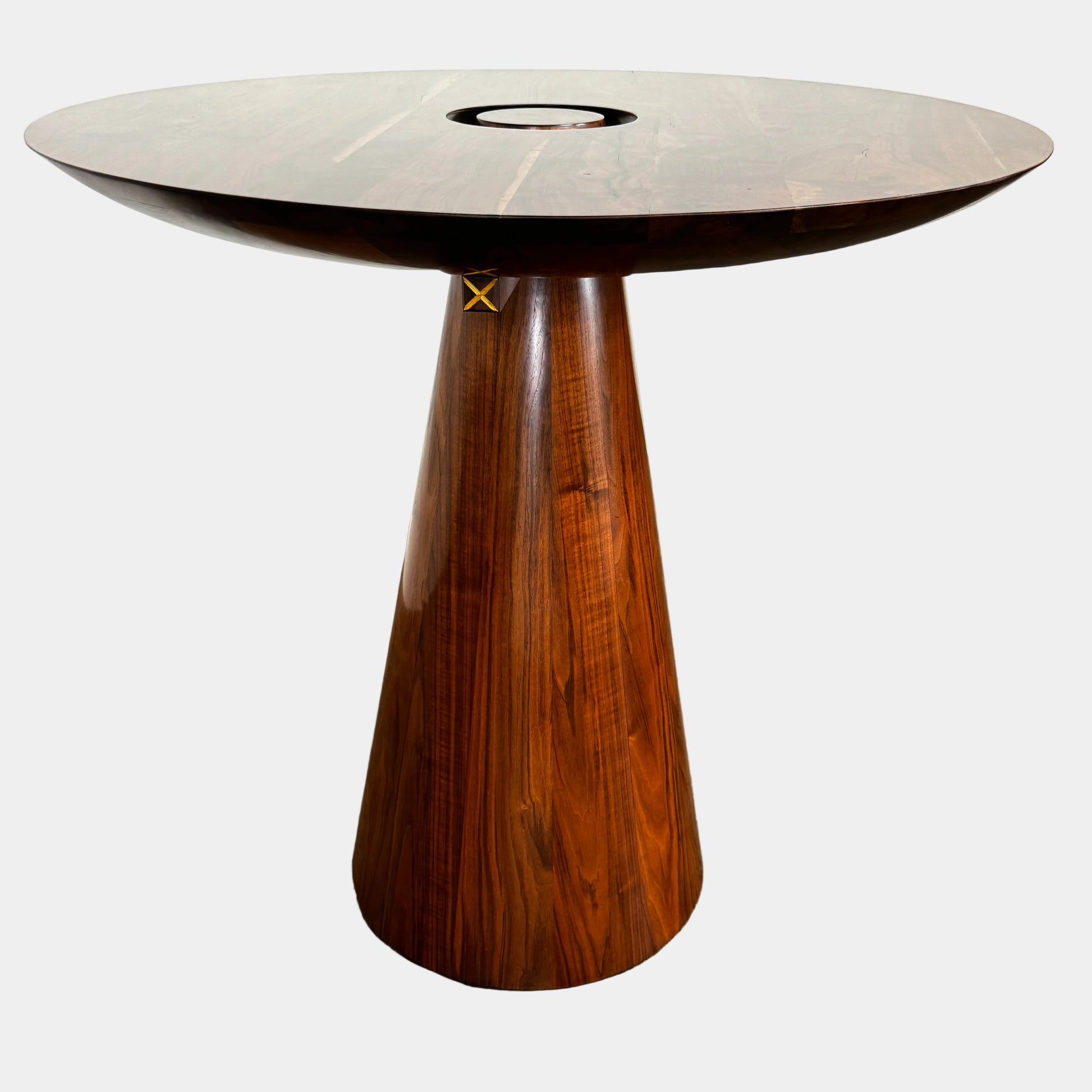 The Mike Fair Claro Walnut Bar Height Table by Artist features a round claro walnut top with a central circular cutout, a conical base, and a small gold cross detail on the side, shown on a concrete floor.