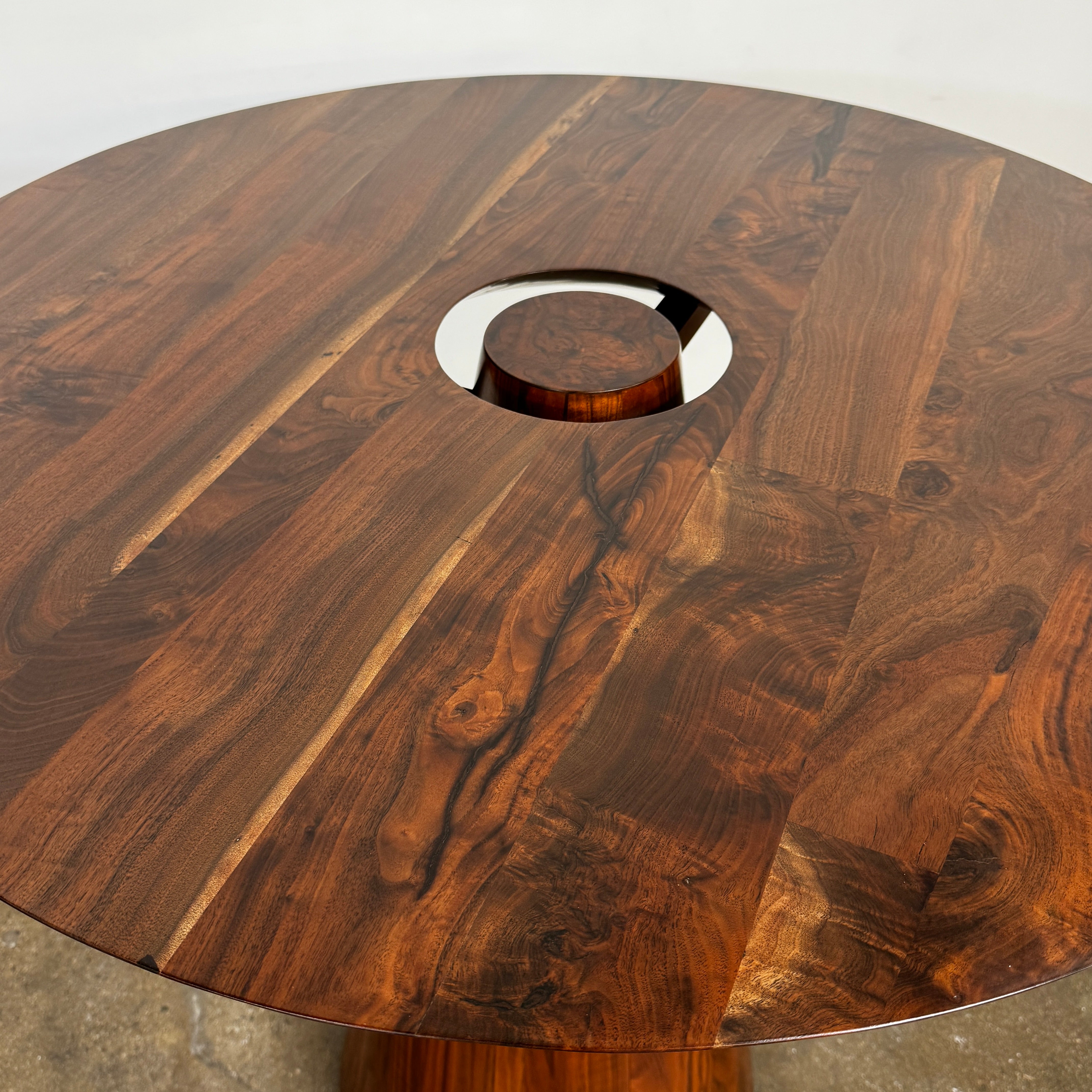 The Mike Fair Claro Walnut Bar Height Table by Artist features a round claro walnut top with a central circular cutout, a conical base, and a small gold cross detail on the side, shown on a concrete floor.