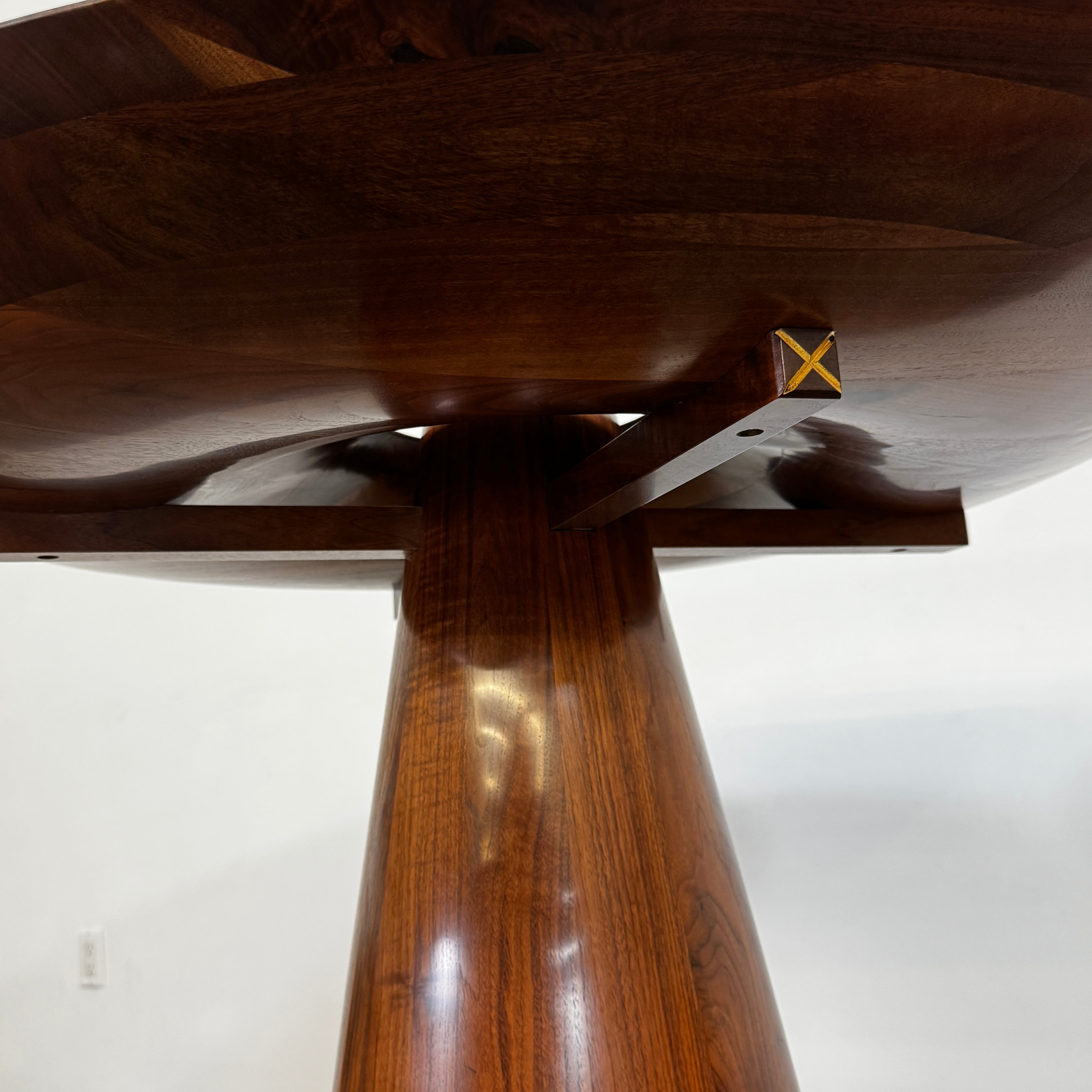 The Mike Fair Claro Walnut Bar Height Table by Artist features a round claro walnut top with a central circular cutout, a conical base, and a small gold cross detail on the side, shown on a concrete floor.