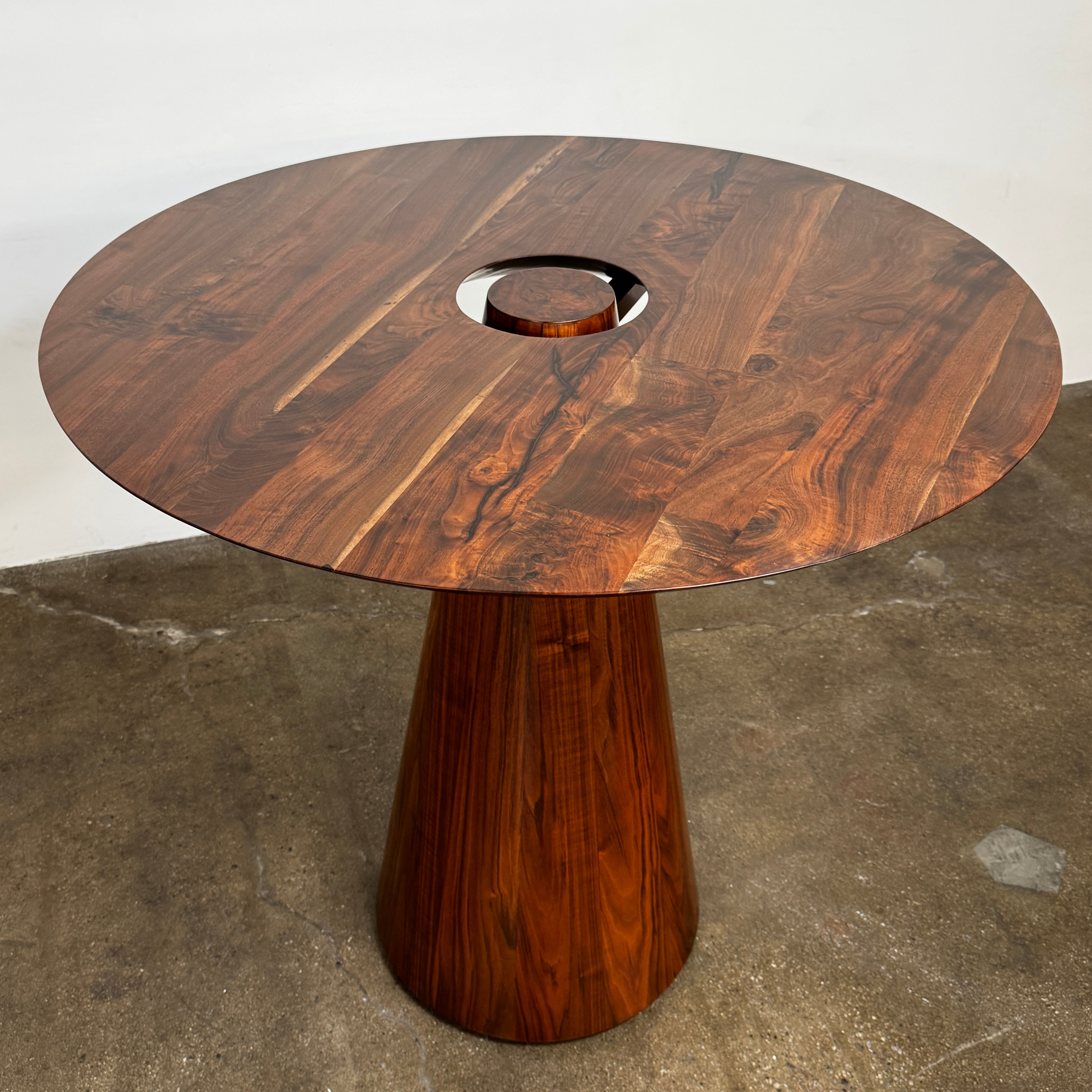 The Mike Fair Claro Walnut Bar Height Table by Artist features a round claro walnut top with a central circular cutout, a conical base, and a small gold cross detail on the side, shown on a concrete floor.