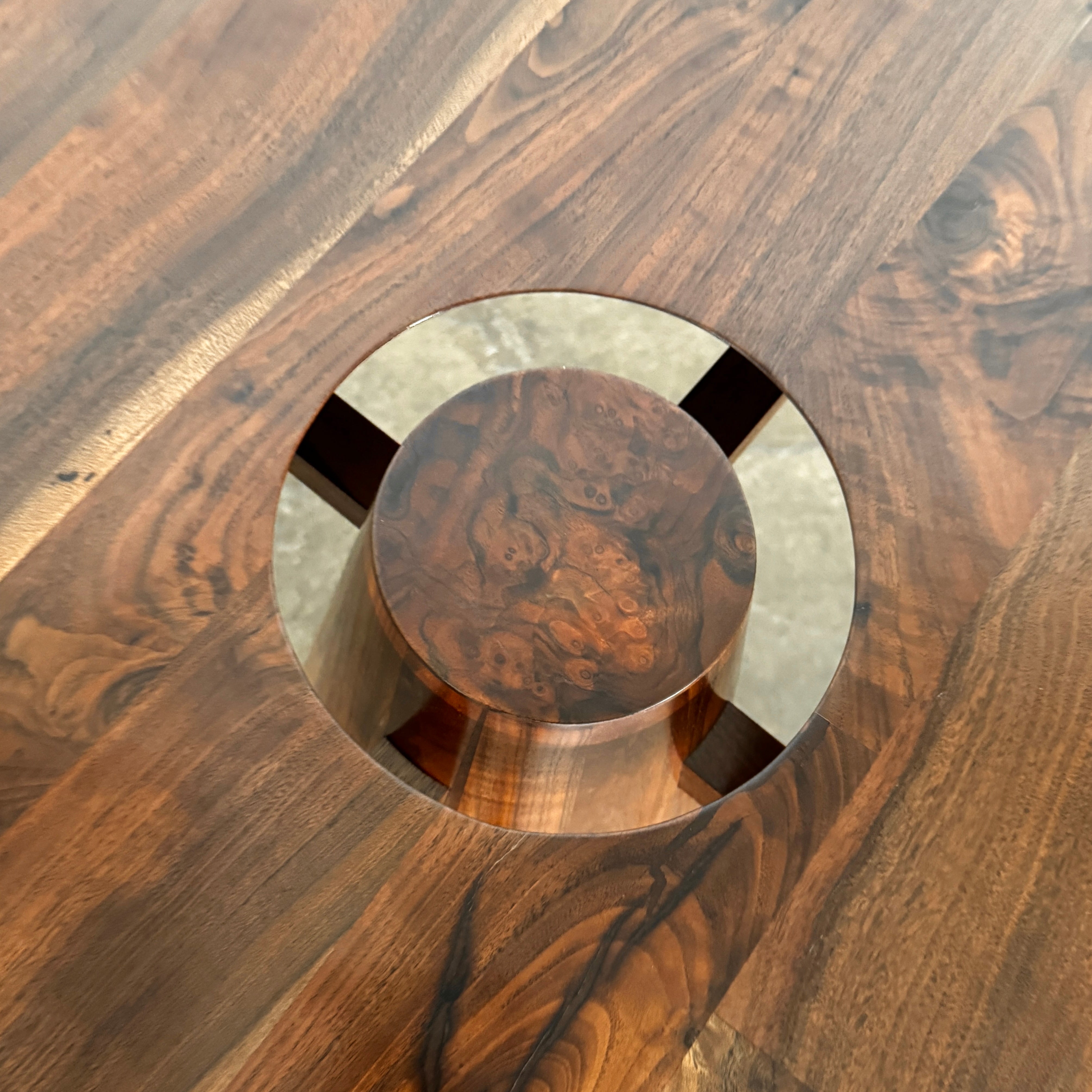 The Mike Fair Claro Walnut Bar Height Table by Artist features a round claro walnut top with a central circular cutout, a conical base, and a small gold cross detail on the side, shown on a concrete floor.