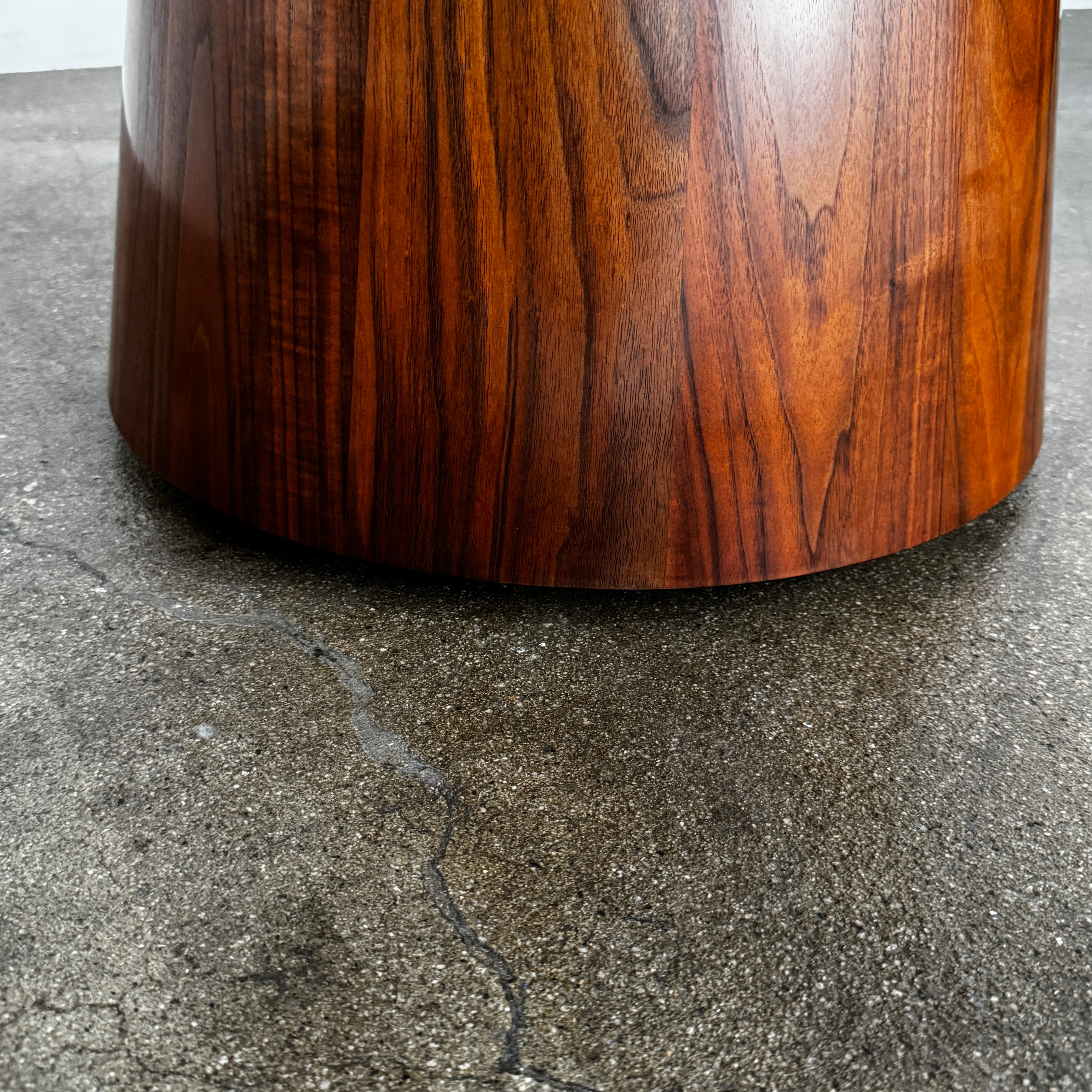 The Mike Fair Claro Walnut Bar Height Table by Artist features a round claro walnut top with a central circular cutout, a conical base, and a small gold cross detail on the side, shown on a concrete floor.