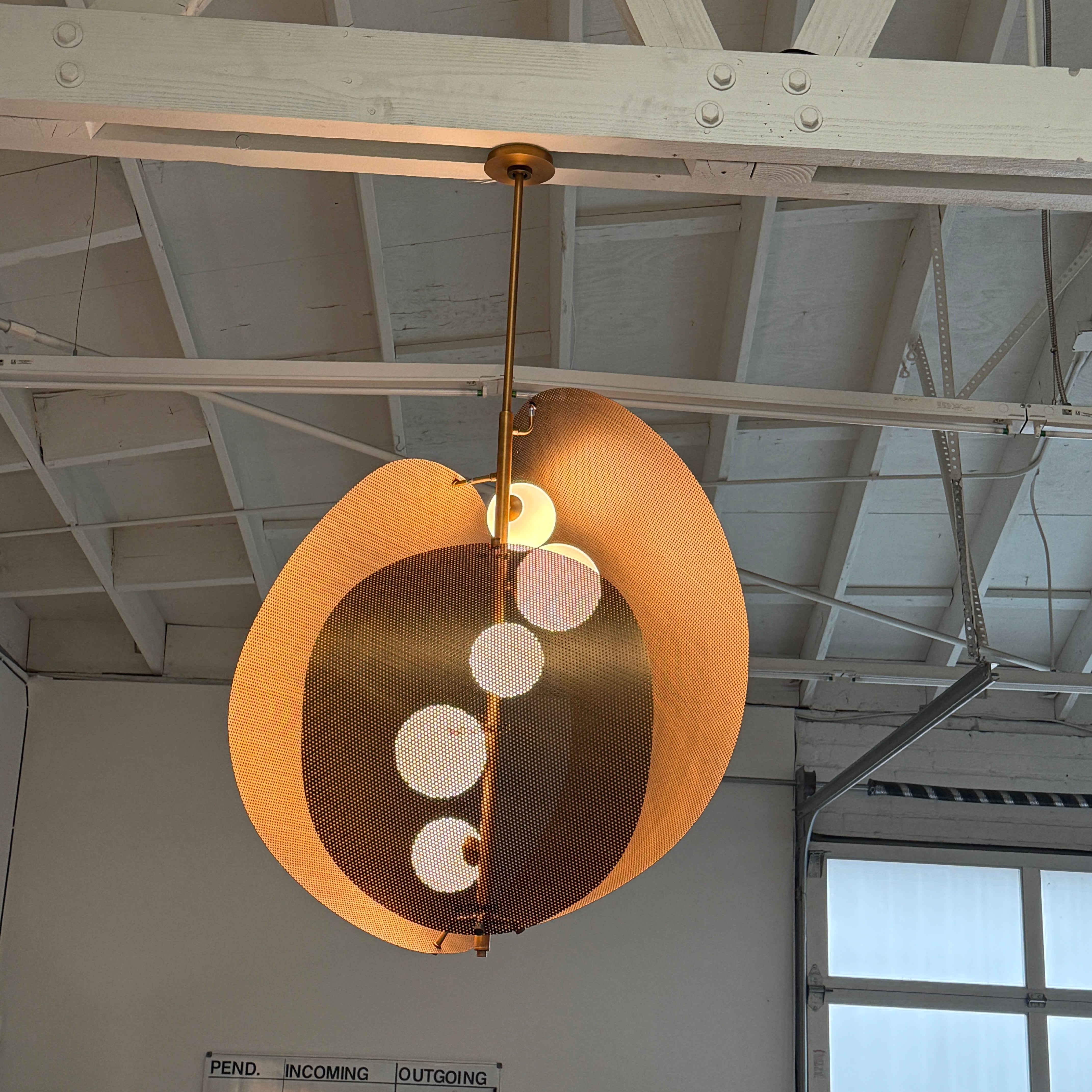 A modern chandelier with circular metal mesh panels and a central bulb hangs from a golden rod, reminiscent of the Studio M Chips 5-Light Vertical Pendant in Natural Aged Brass.