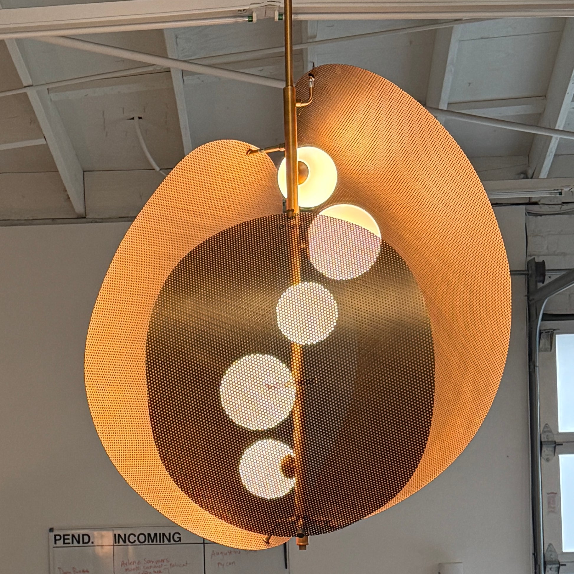 A modern chandelier with circular metal mesh panels and a central bulb hangs from a golden rod, reminiscent of the Studio M Chips 5-Light Vertical Pendant in Natural Aged Brass.
