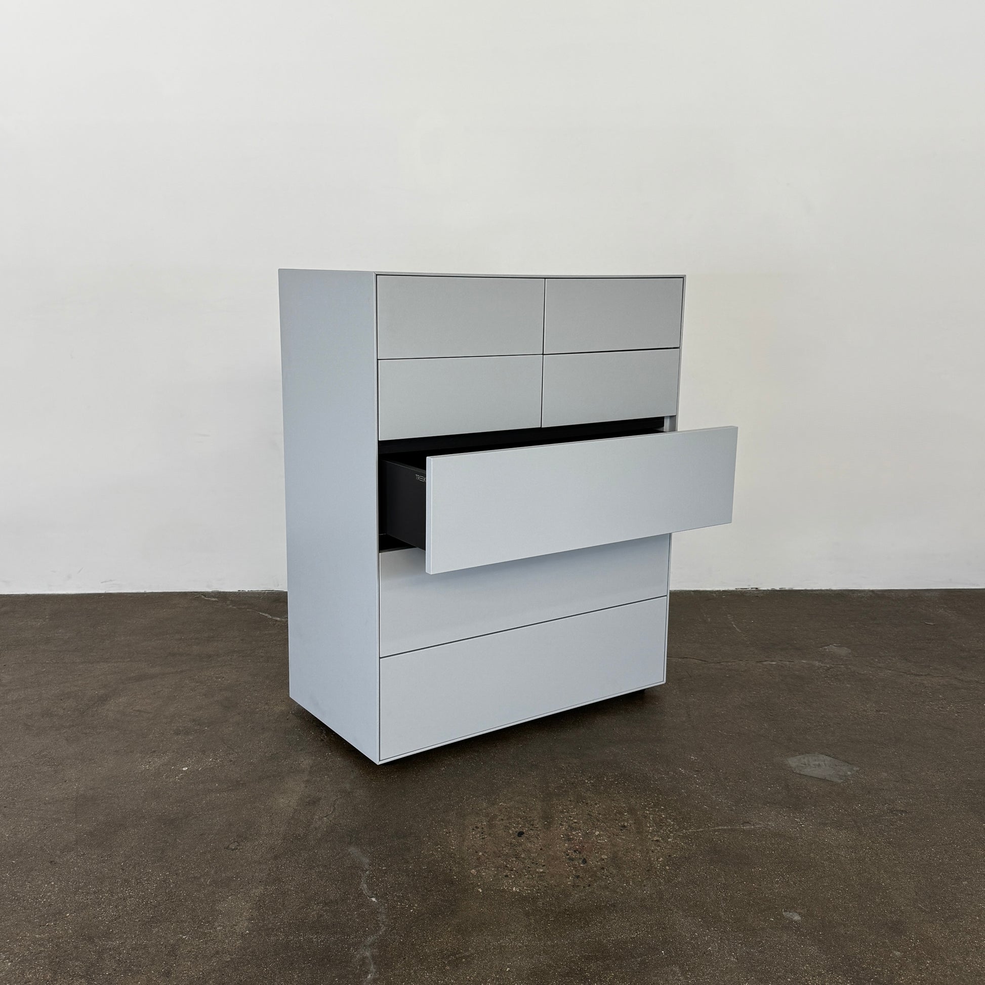 The Treku Lauki Tall Dresser by Treku, with a sleek lacquered finish and push-latch drawers—one partially open—stands on a concrete floor against a plain white wall.