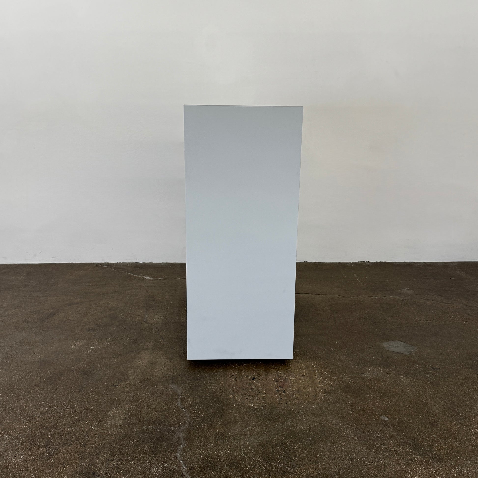 The Treku Lauki Tall Dresser by Treku, with a sleek lacquered finish and push-latch drawers—one partially open—stands on a concrete floor against a plain white wall.