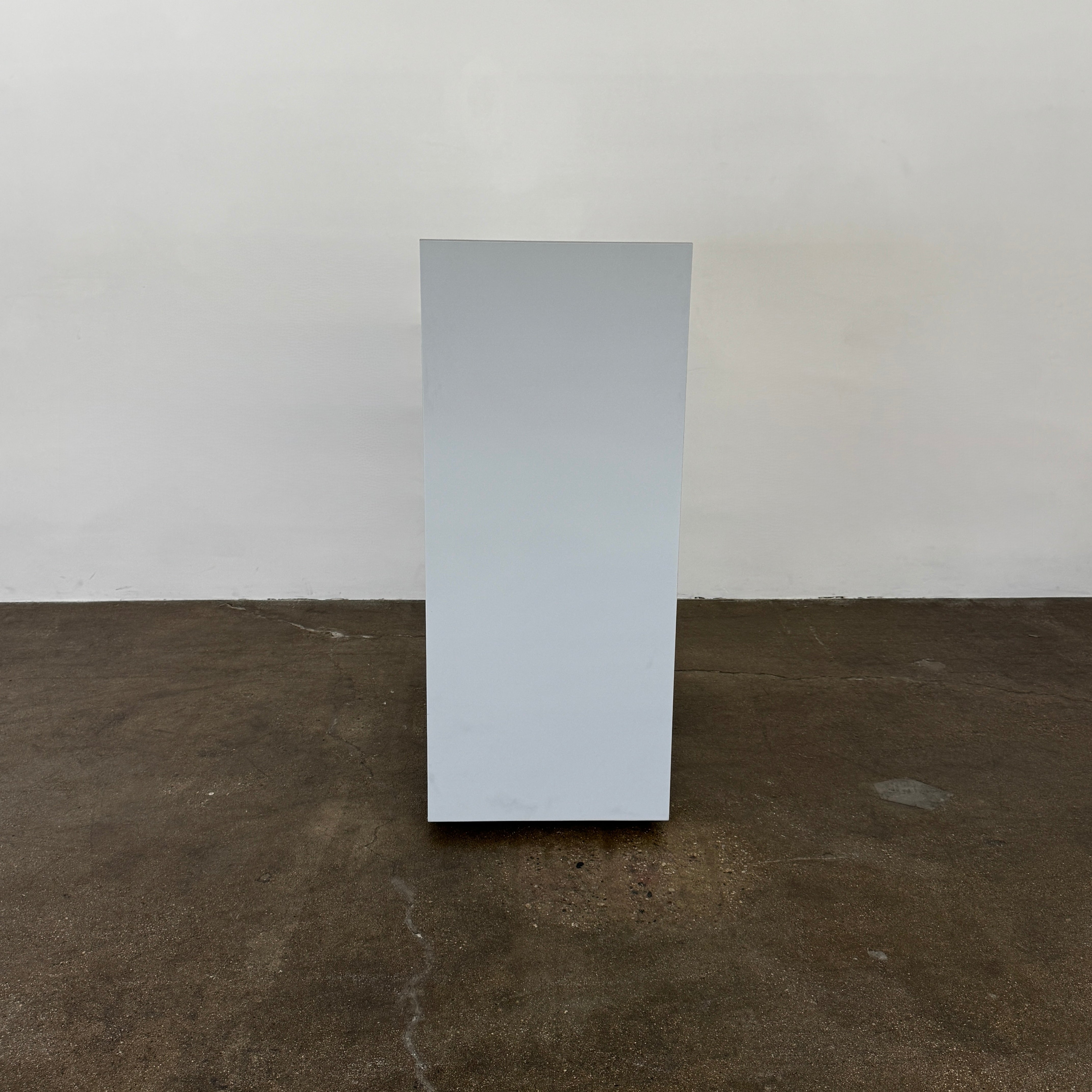 The Treku Lauki Tall Dresser by Treku, with a sleek lacquered finish and push-latch drawers—one partially open—stands on a concrete floor against a plain white wall.