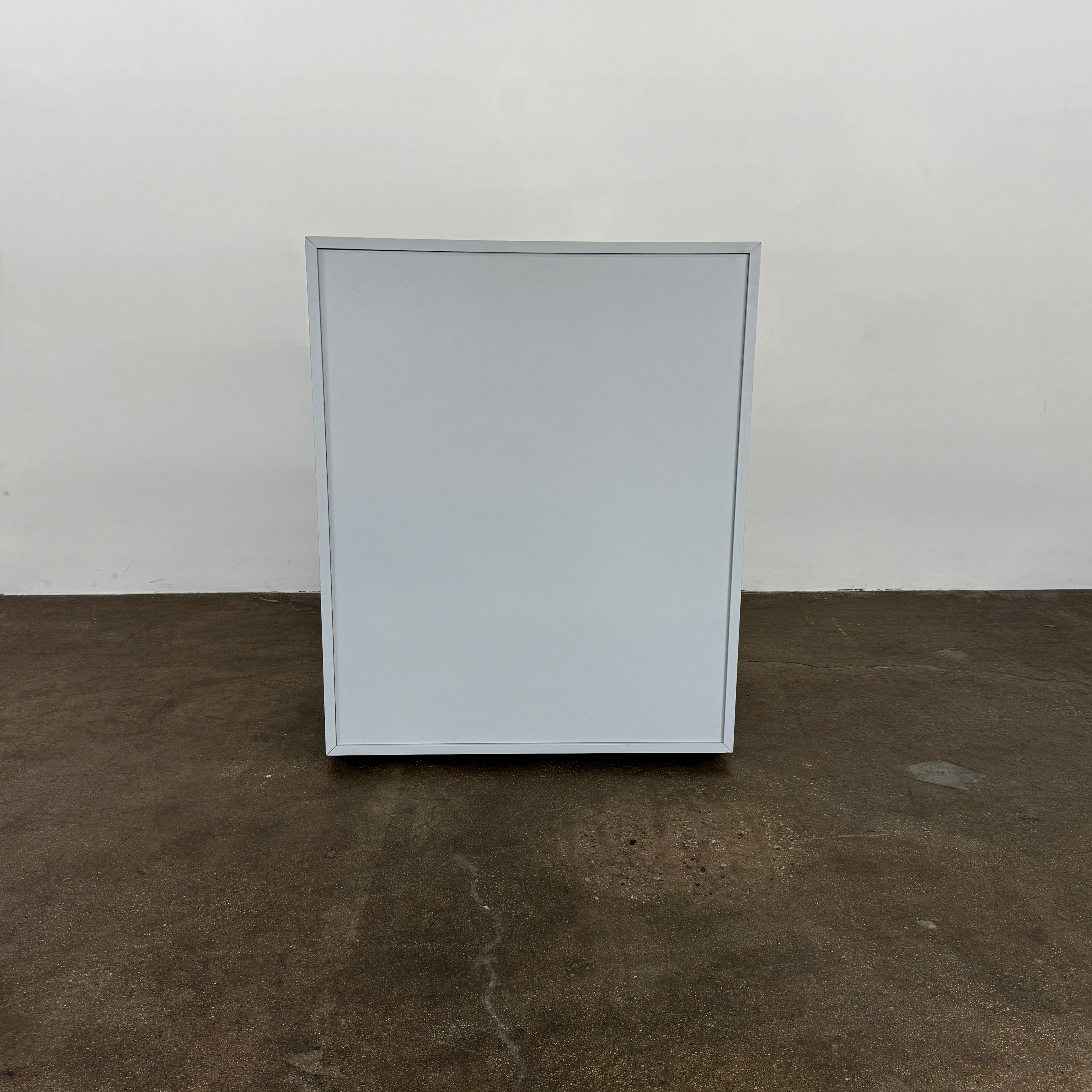 The Treku Lauki Tall Dresser by Treku, with a sleek lacquered finish and push-latch drawers—one partially open—stands on a concrete floor against a plain white wall.