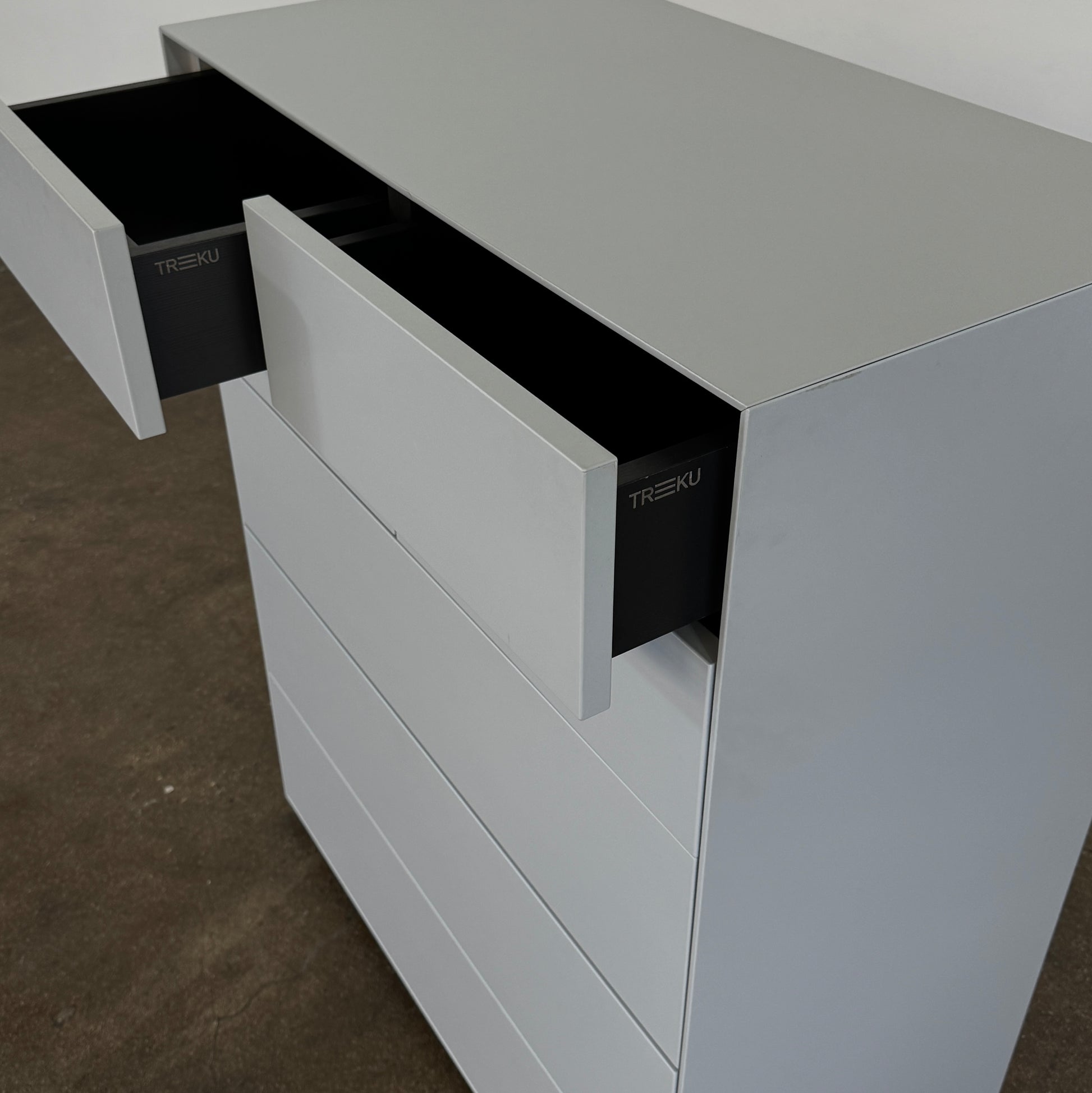 The Treku Lauki Tall Dresser by Treku, with a sleek lacquered finish and push-latch drawers—one partially open—stands on a concrete floor against a plain white wall.