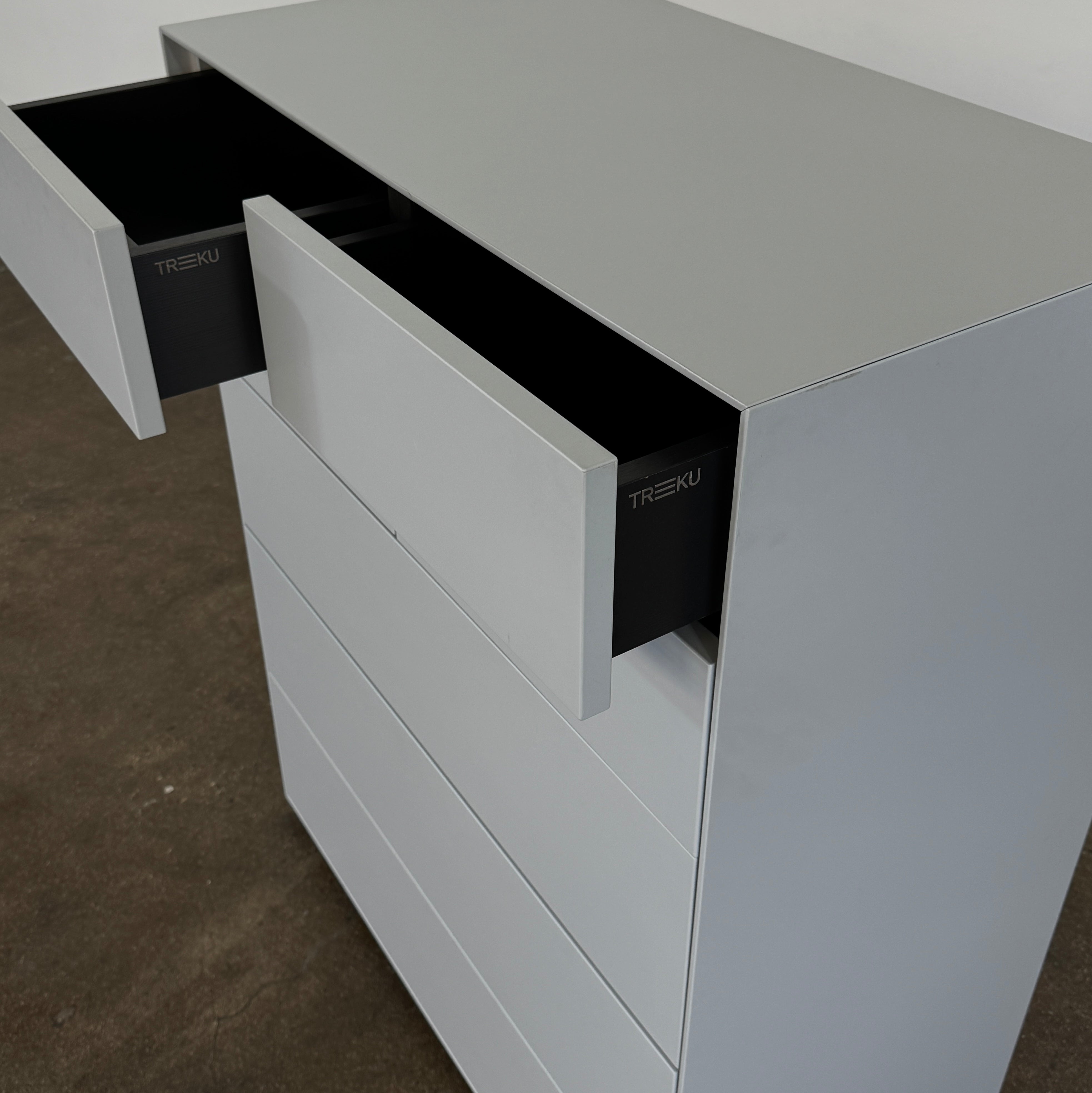 The Treku Lauki Tall Dresser by Treku, with a sleek lacquered finish and push-latch drawers—one partially open—stands on a concrete floor against a plain white wall.