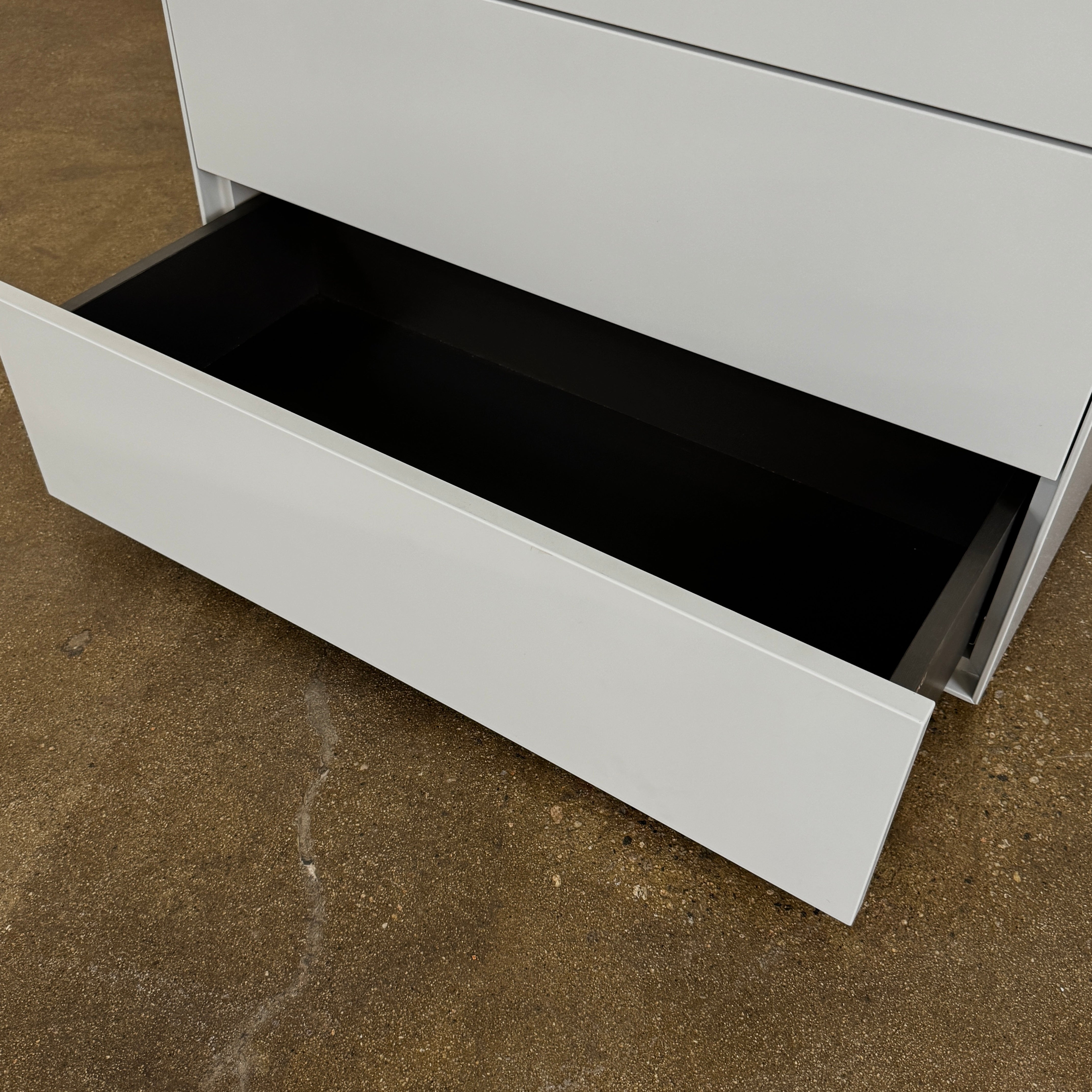 The Treku Lauki Tall Dresser by Treku, with a sleek lacquered finish and push-latch drawers—one partially open—stands on a concrete floor against a plain white wall.