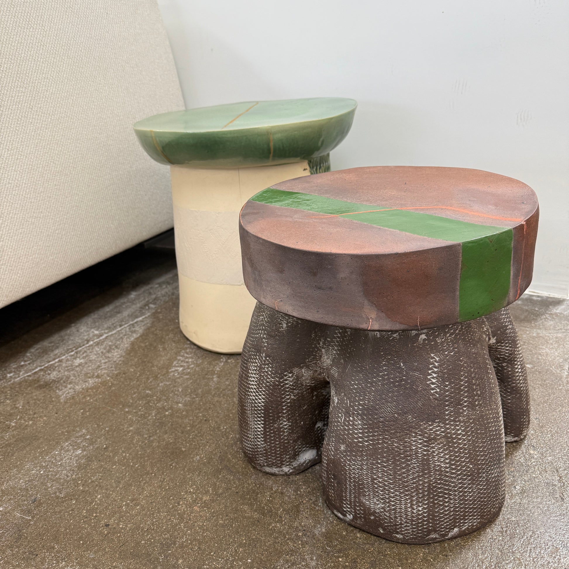 The Allyn Davis Mrs Beastie Ceramic Side Table by Artist features a round, textured design with a wooden top and green stripe. Its woven four-legged base subtly incorporates Kintsugo elements, adding depth and character to the piece.
