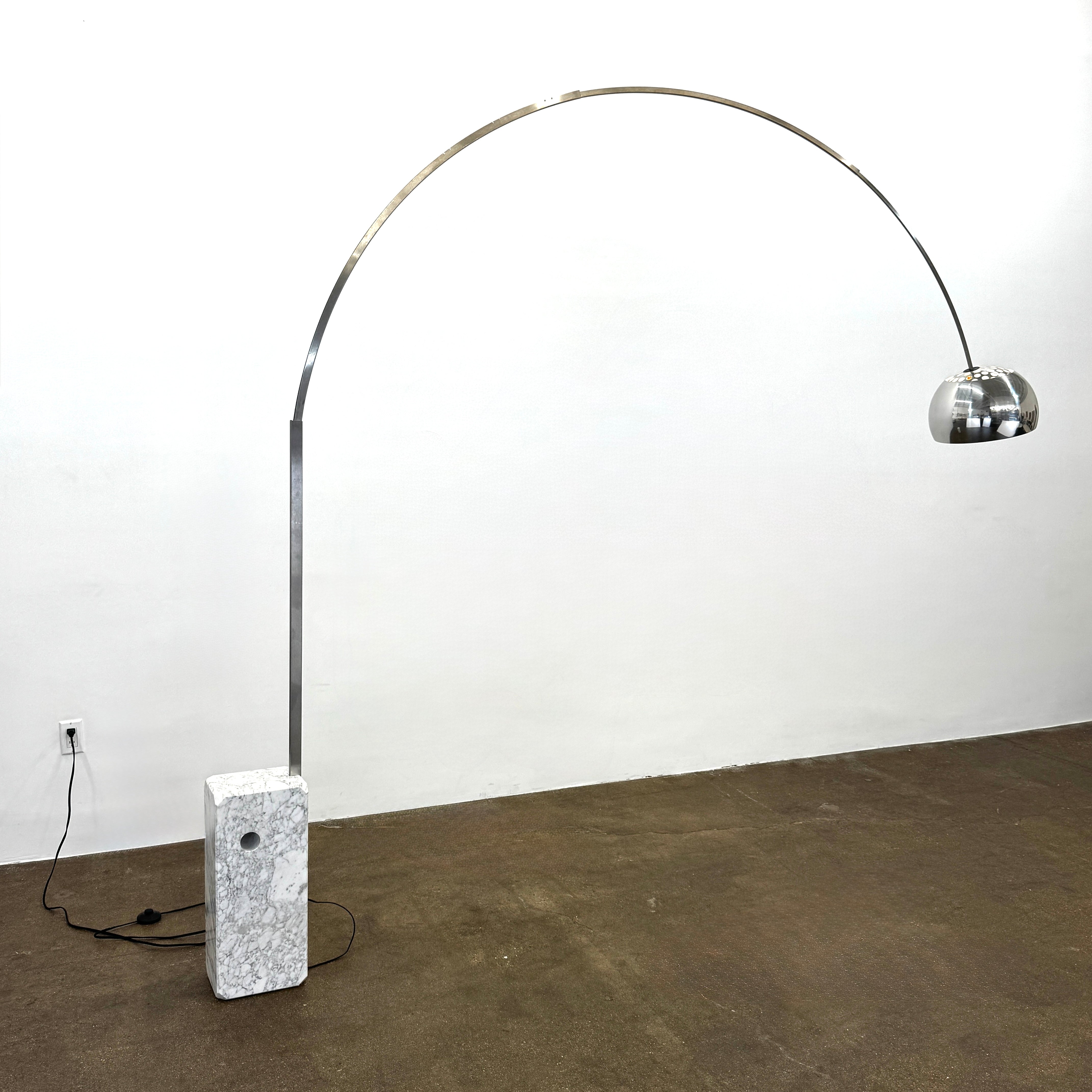 The Flos Arco Floor Lamp by Flos features a curved metal arm, dome-shaped metallic shade, and a rectangular marble base, inspired by the iconic original design.