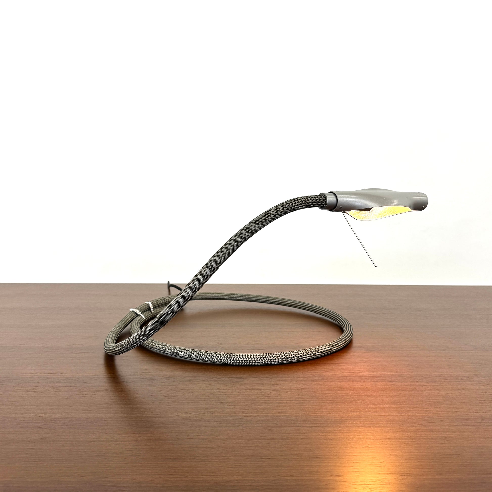 The Ingo Maurer Metall T. Cooper Task Light features a flexible metal braided hose with a metallic nozzle on one end and exposed wires on the other, coiled on a plain white background—evoking collectible German designer lighting.