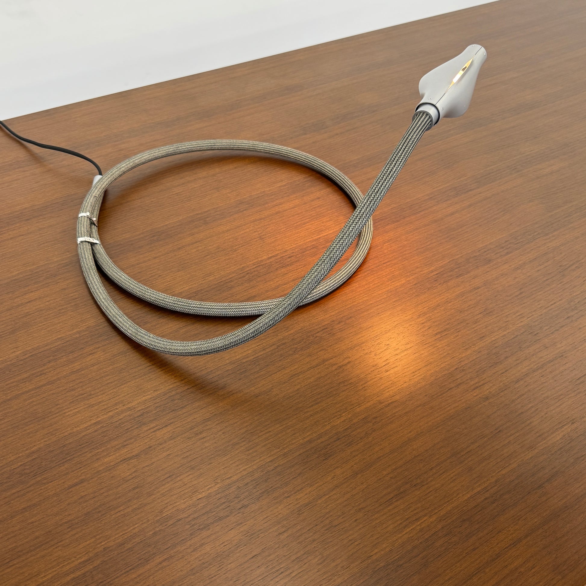 The Ingo Maurer Metall T. Cooper Task Light features a flexible metal braided hose with a metallic nozzle on one end and exposed wires on the other, coiled on a plain white background—evoking collectible German designer lighting.