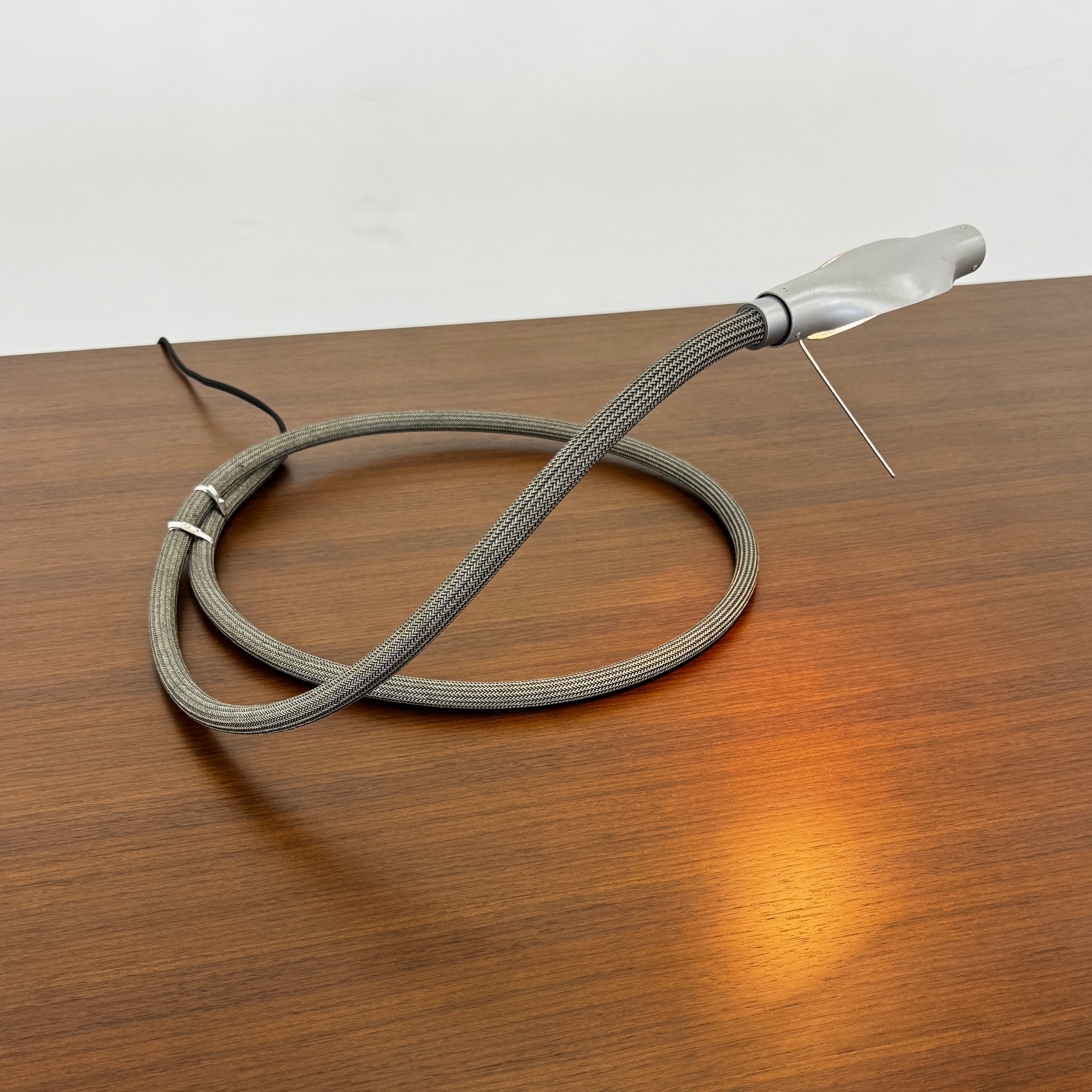 The Ingo Maurer Metall T. Cooper Task Light features a flexible metal braided hose with a metallic nozzle on one end and exposed wires on the other, coiled on a plain white background—evoking collectible German designer lighting.