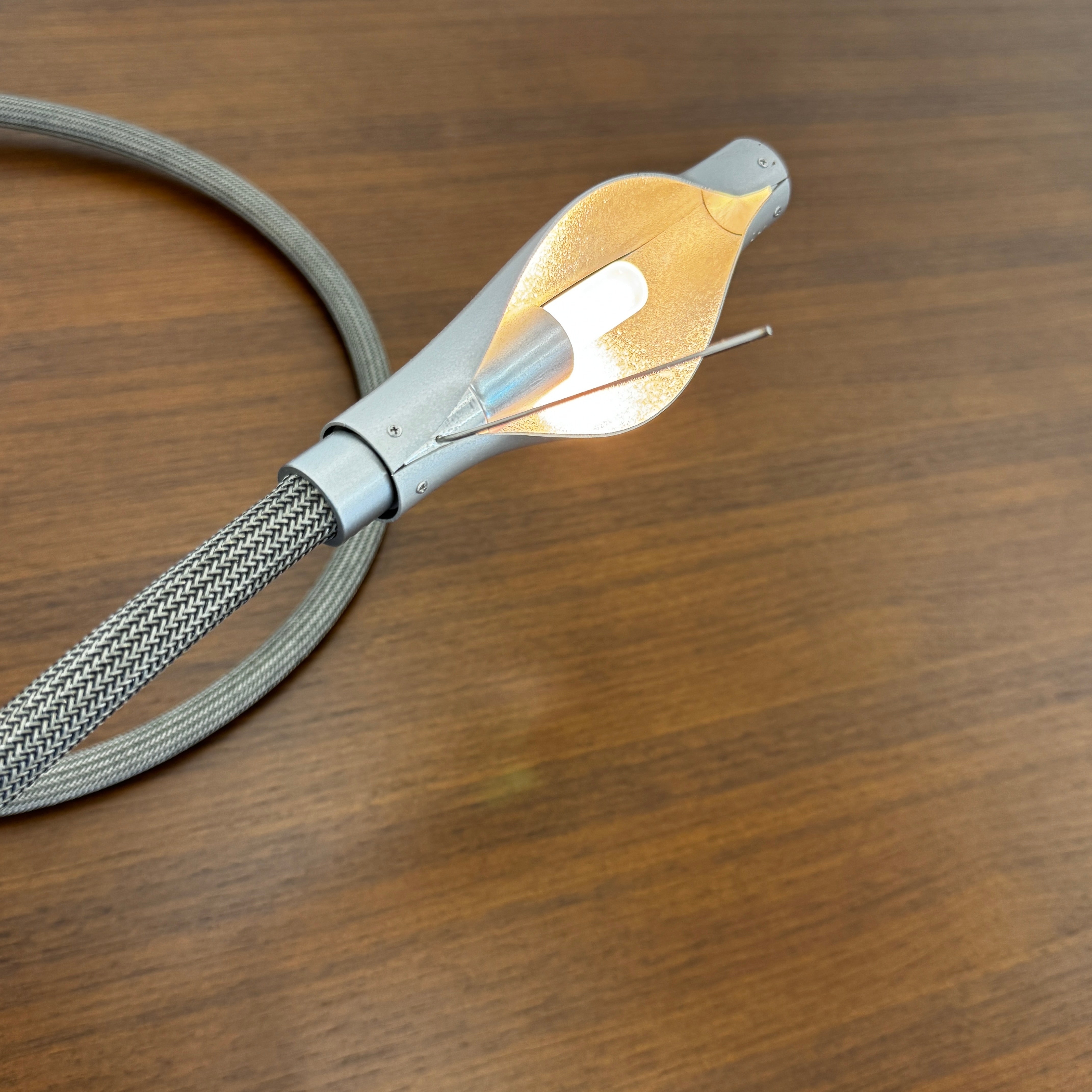 The Ingo Maurer Metall T. Cooper Task Light features a flexible metal braided hose with a metallic nozzle on one end and exposed wires on the other, coiled on a plain white background—evoking collectible German designer lighting.