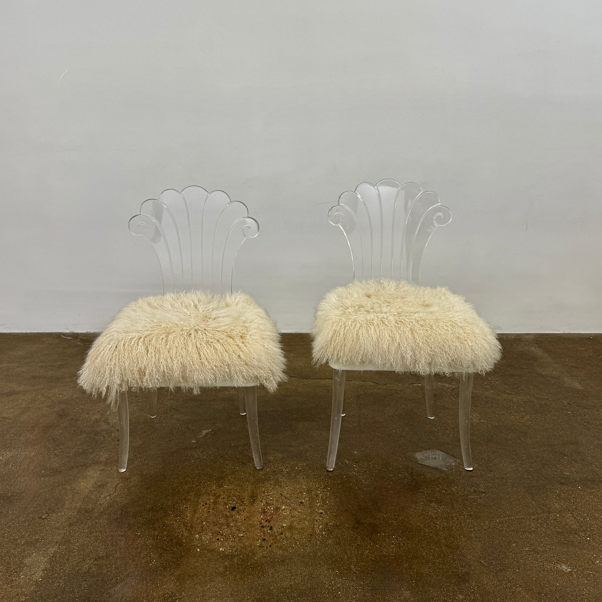 Two white Vintage Elsie de Wolfe Lucite Chairs with elegant fan-shaped backs and plush beige sheepskin seats are displayed against a plain white background.