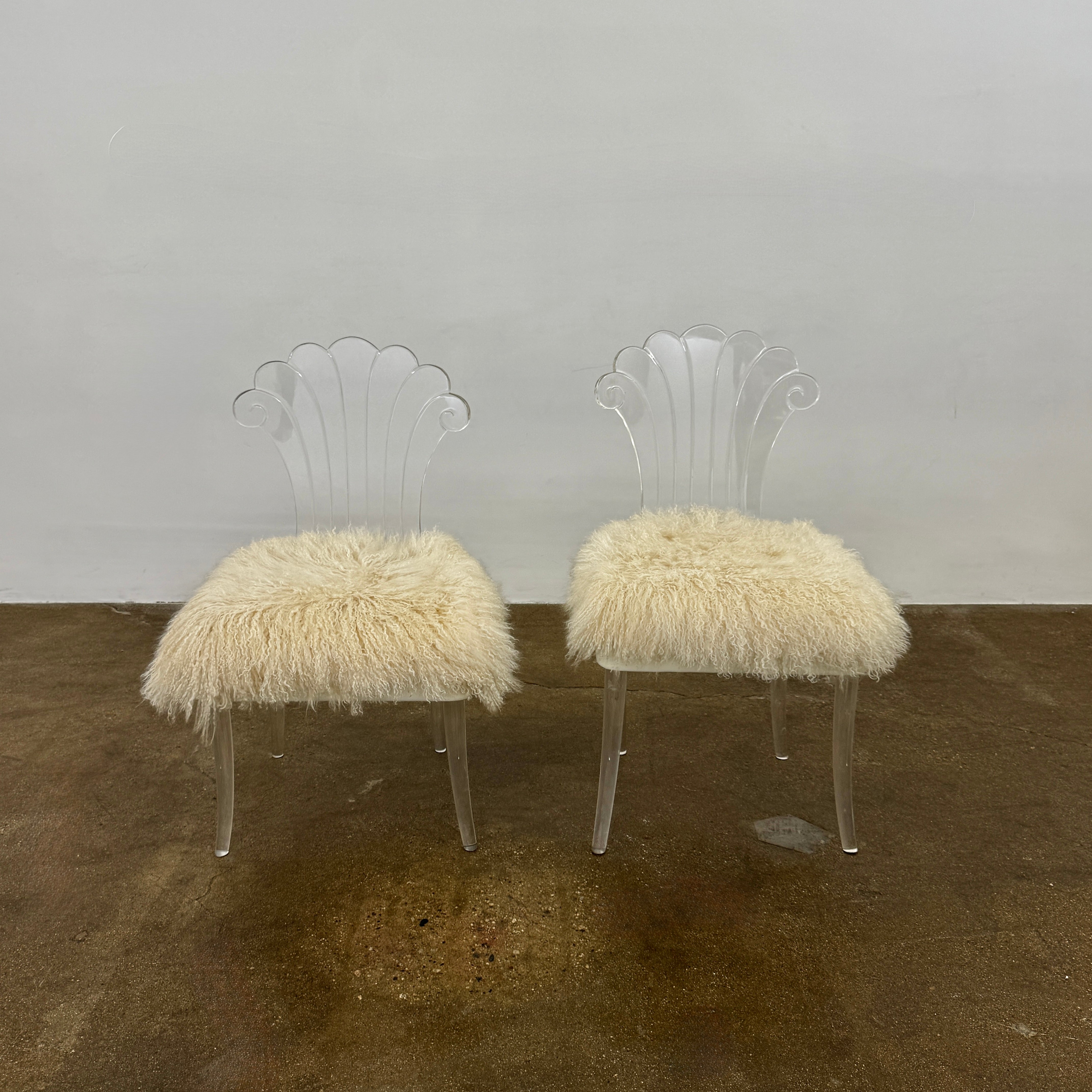 Two white Vintage Elsie de Wolfe Lucite Chairs with elegant fan-shaped backs and plush beige sheepskin seats are displayed against a plain white background.