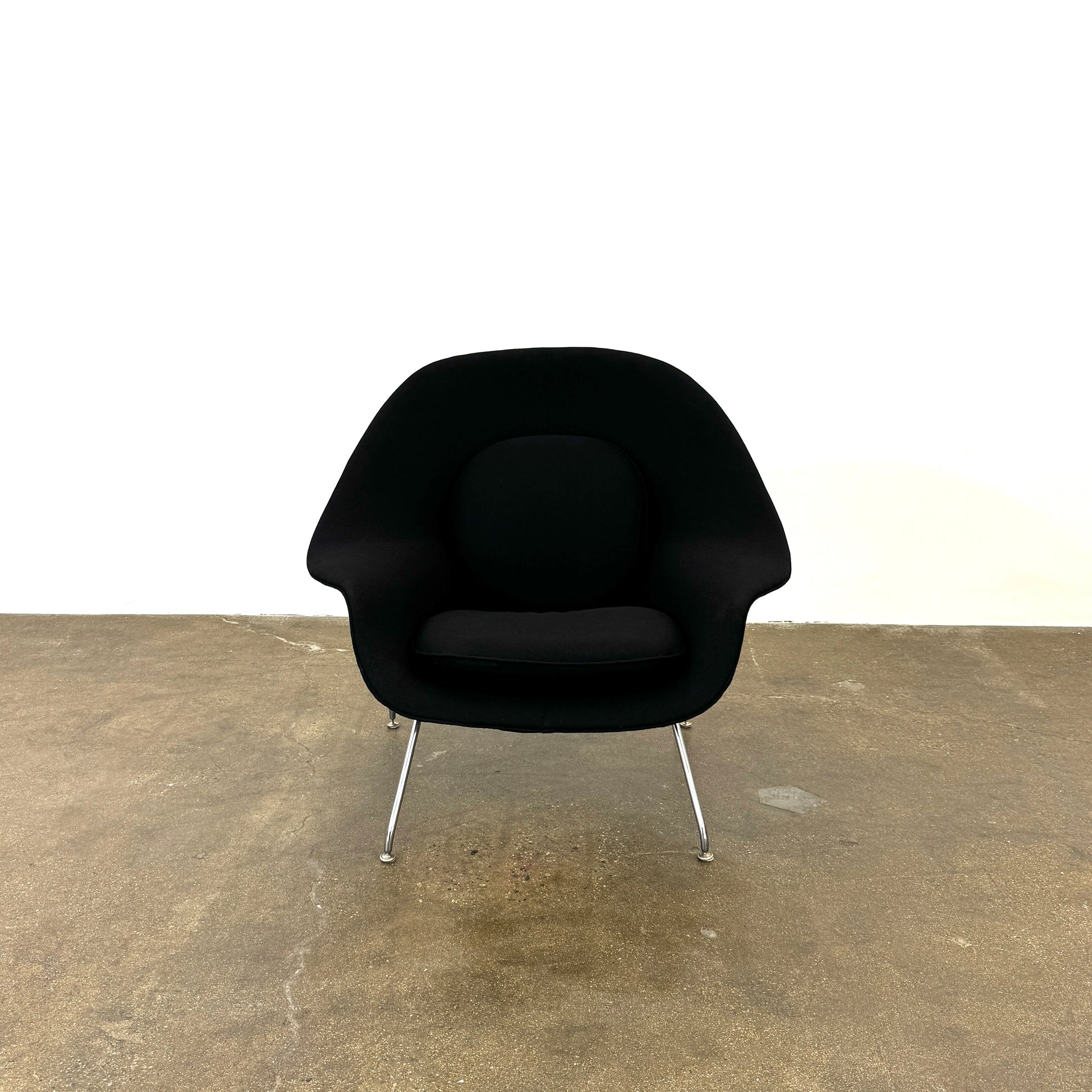 The Knoll Womb Chair by Knoll features a black curved seat, padded cushion, and metal legs, drawing inspiration from midcentury modern design and the iconic Saarinen style. Shown here against a plain white background.