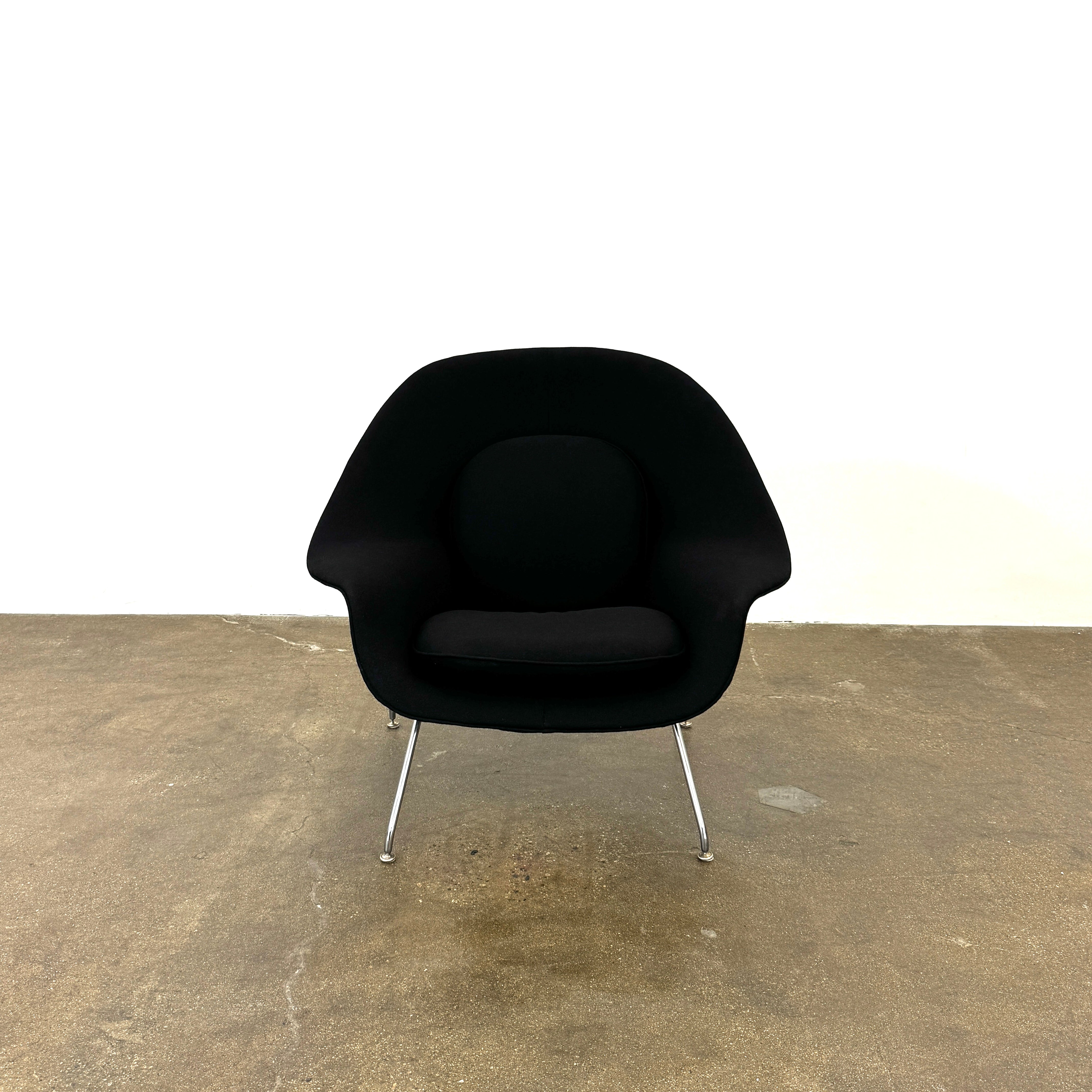 The Knoll Womb Chair by Knoll features a black curved seat, padded cushion, and metal legs, drawing inspiration from midcentury modern design and the iconic Saarinen style. Shown here against a plain white background.