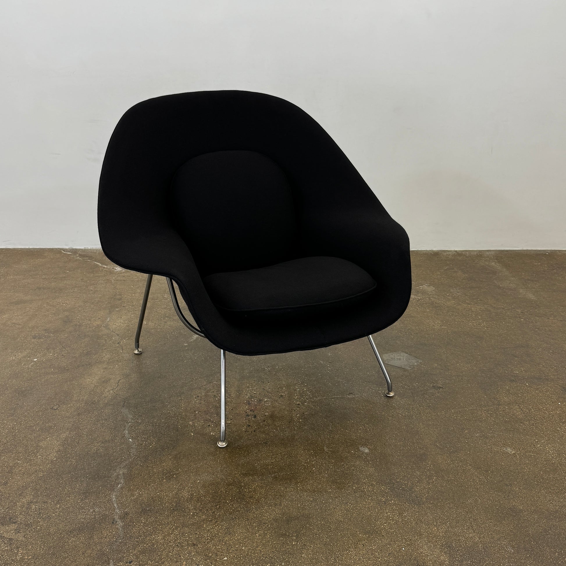 The Knoll Womb Chair by Knoll features a black curved seat, padded cushion, and metal legs, drawing inspiration from midcentury modern design and the iconic Saarinen style. Shown here against a plain white background.