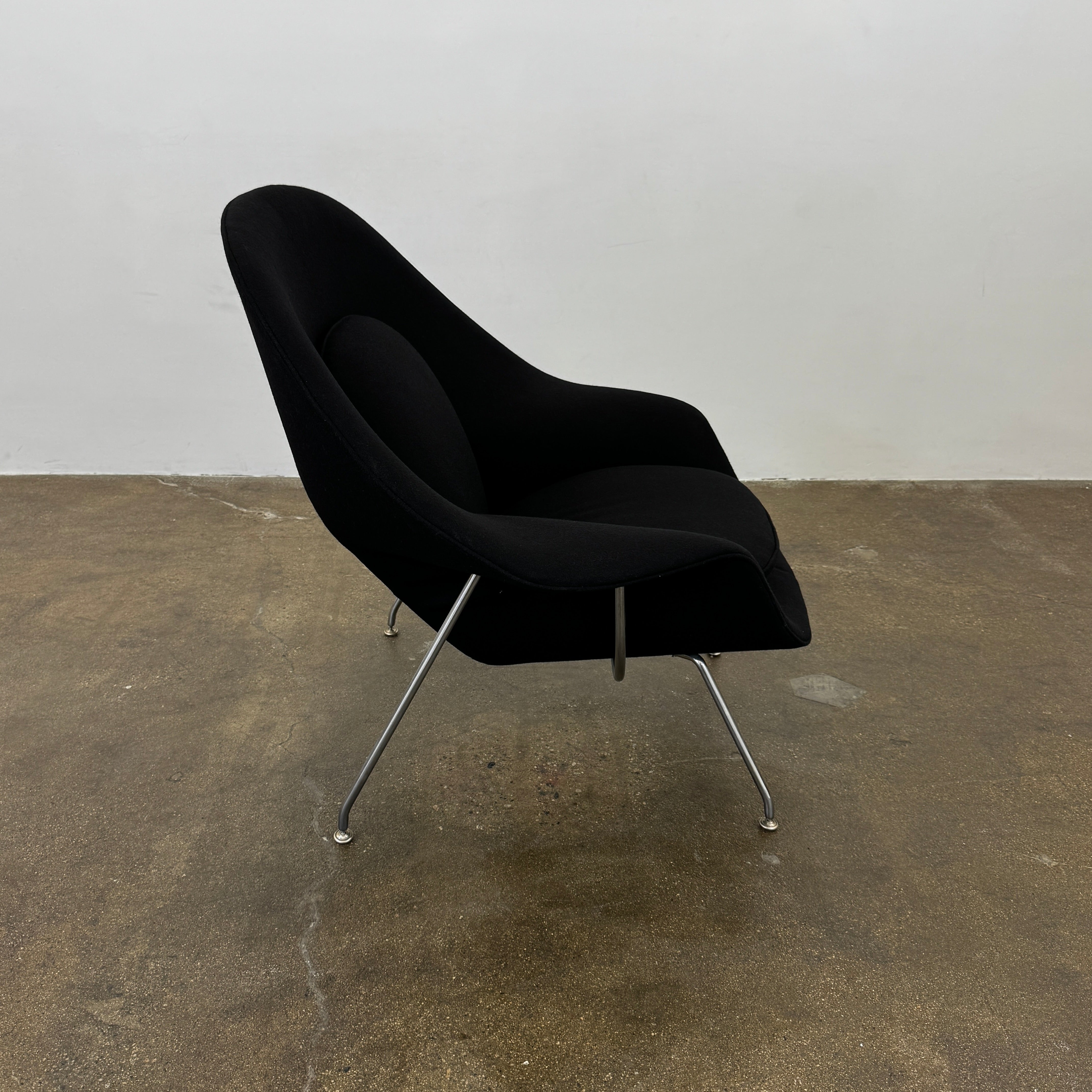 The Knoll Womb Chair by Knoll features a black curved seat, padded cushion, and metal legs, drawing inspiration from midcentury modern design and the iconic Saarinen style. Shown here against a plain white background.