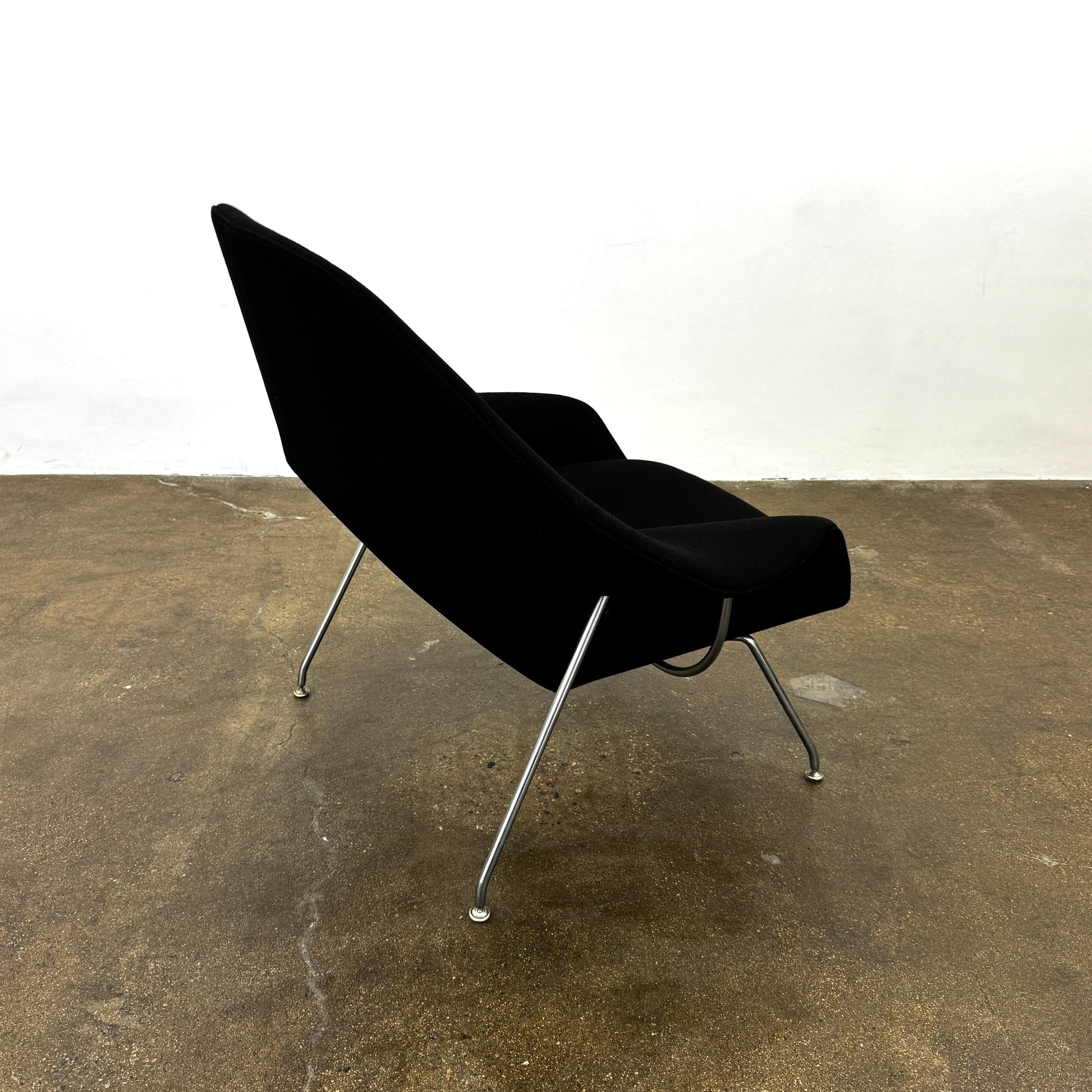 The Knoll Womb Chair by Knoll features a black curved seat, padded cushion, and metal legs, drawing inspiration from midcentury modern design and the iconic Saarinen style. Shown here against a plain white background.