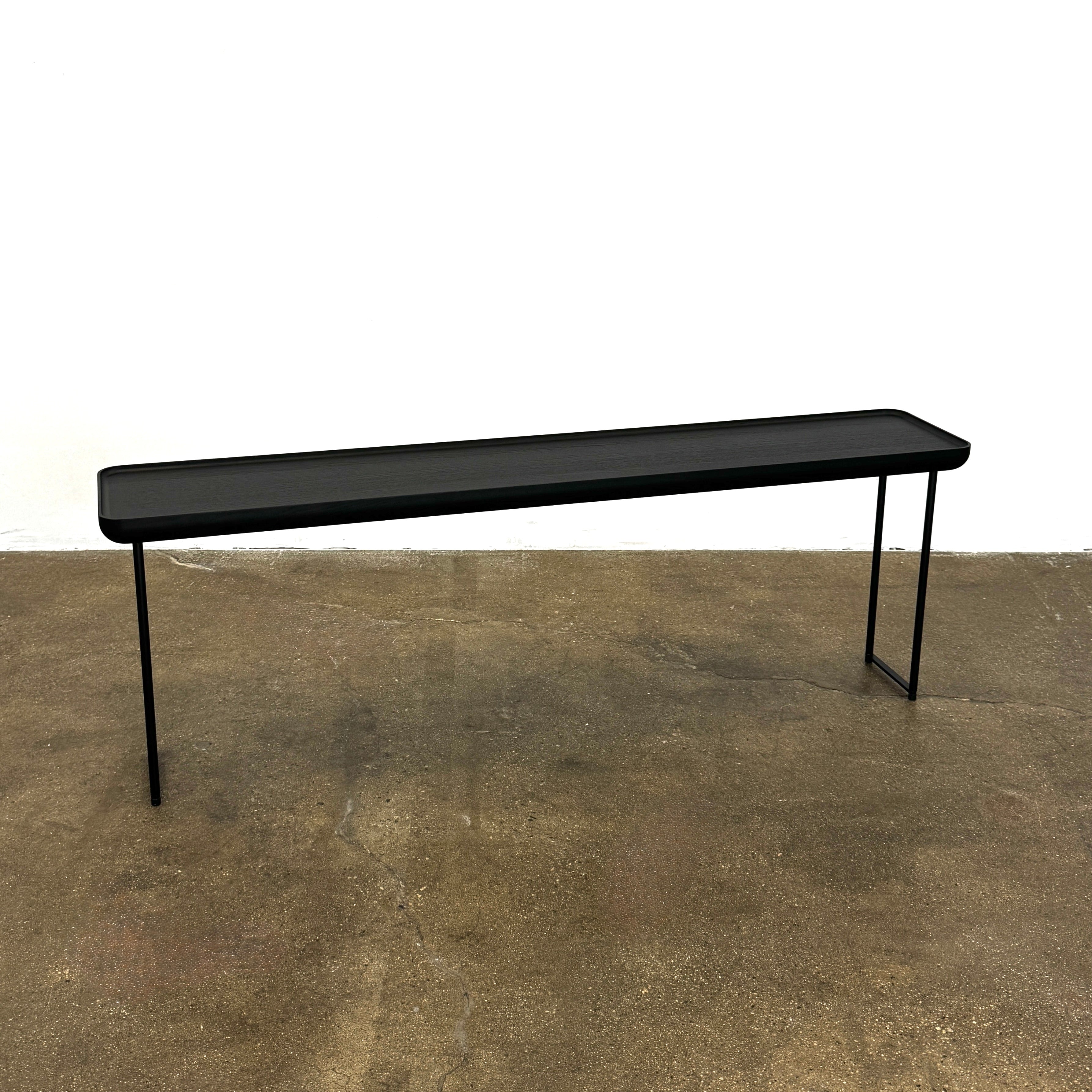 A minimalist black bench with a rectangular seat and slim metal legs echoes the elegant design of the Cassina Torei Occasional Table in Black Ash by Cassina, displayed against a plain white background.