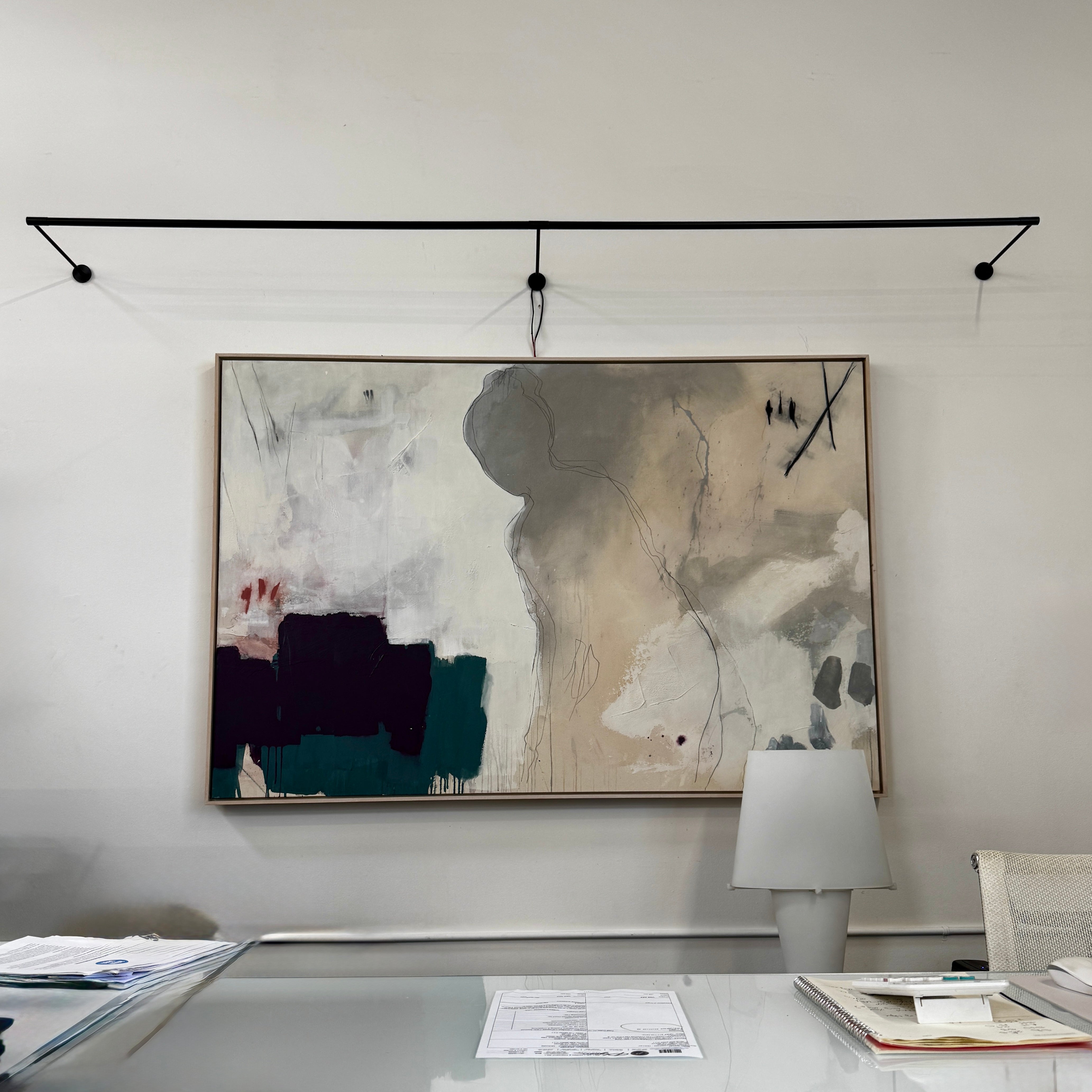 A modern abstract painting hangs on a white wall above a desk, illuminated by the Juniper THIN System Linear Light Wall-mounted from Juniper, with a lamp, documents, and office supplies arranged on the glass desktop.