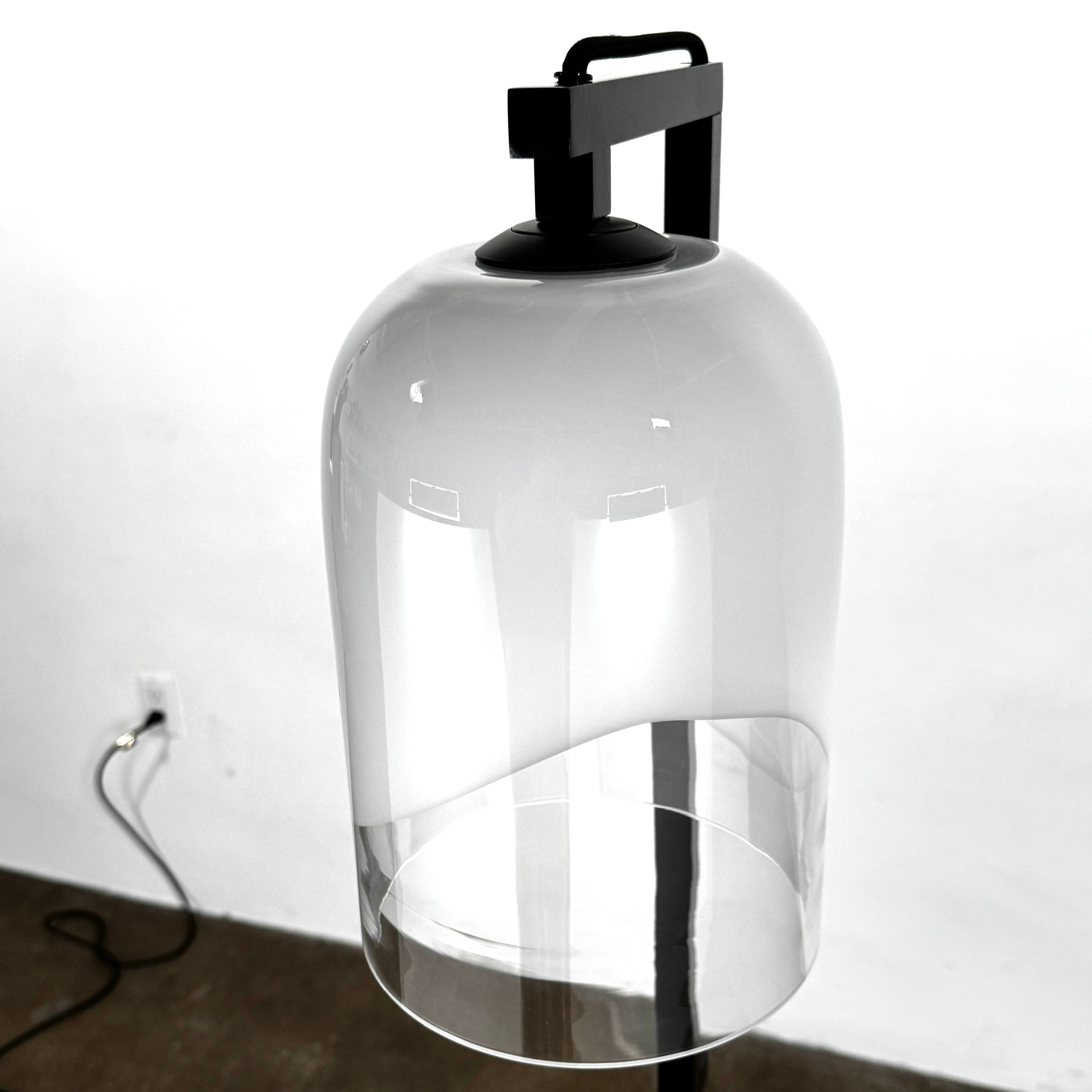 The Articolo Moni Floor Lamp by Articolo features a modern, artisanal design with a curved mouth-blown glass shade in frosted white and clear sections, shown against a plain light-colored wall.