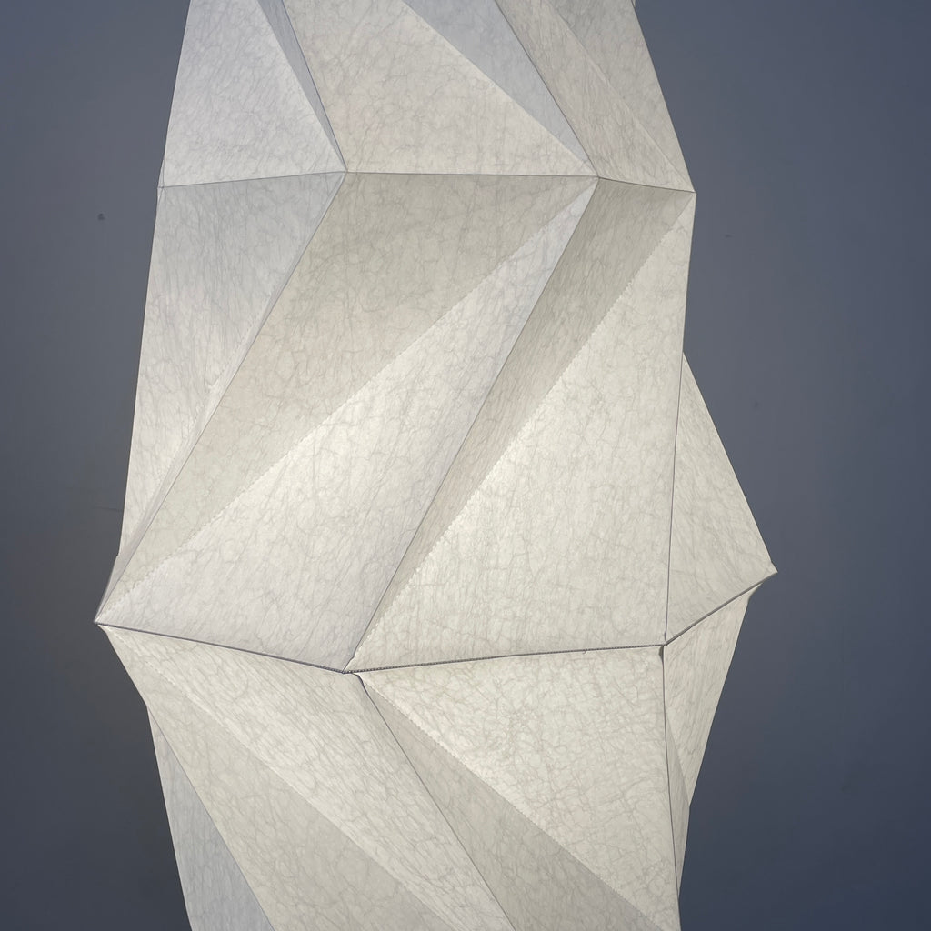 A tall geometric sculpture made of interlocking white panels stands on a simple base, functioning as an Artemide Minomushi floor light in an indoor setting with a concrete floor.