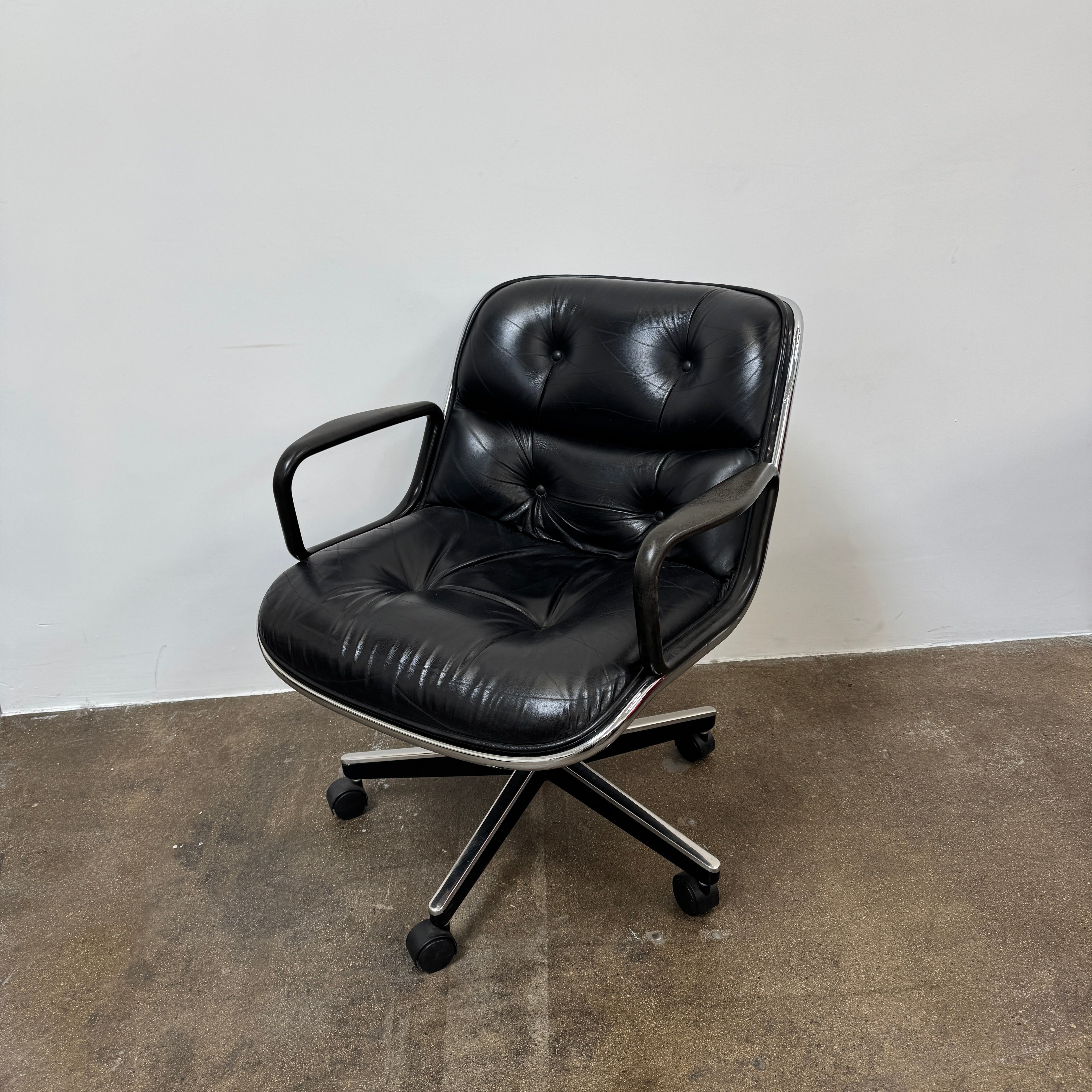 A DWR Pollock Executive Chair with a chrome frame and five wheels is shown from the back, set on a concrete floor against a plain white wall.