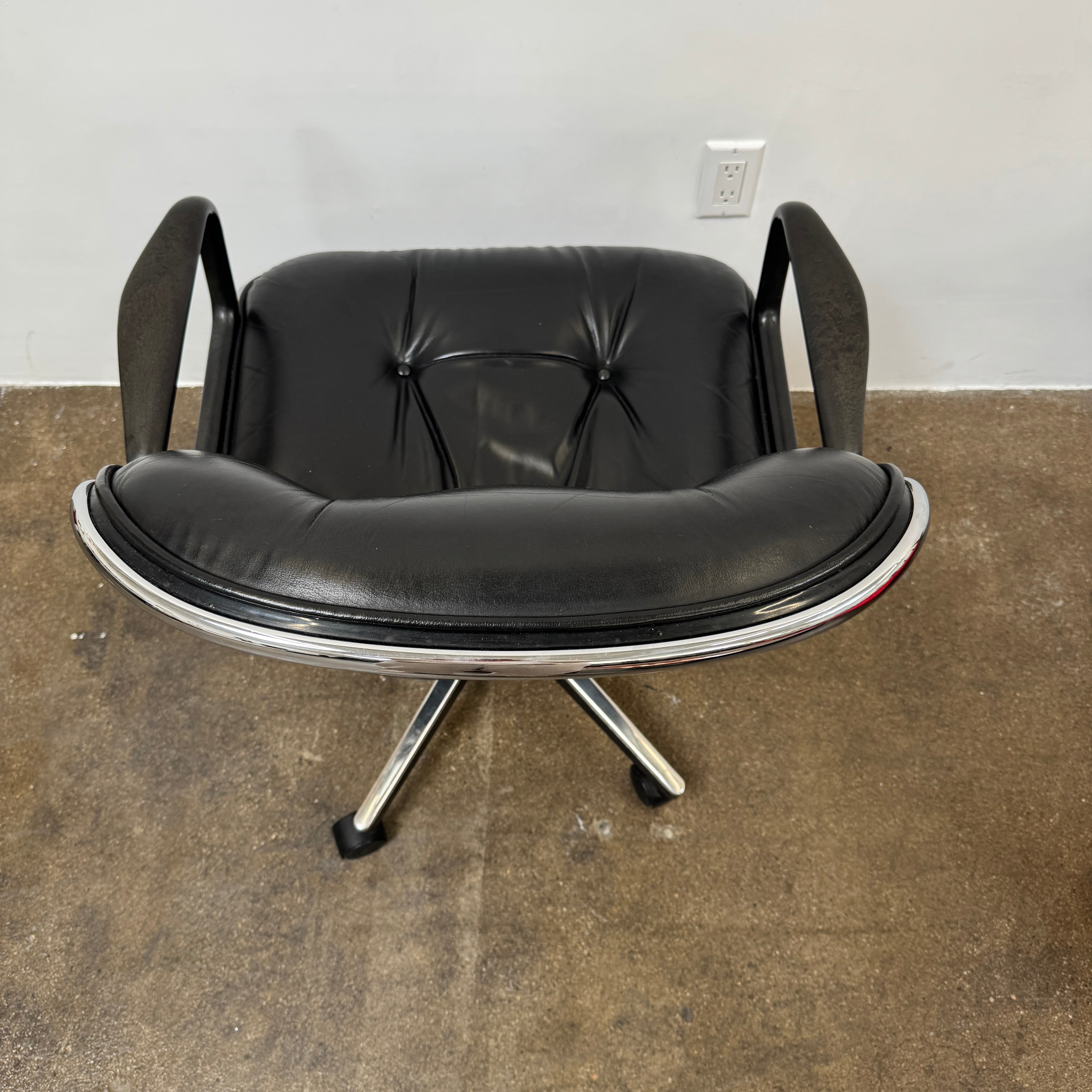 A DWR Pollock Executive Chair with a chrome frame and five wheels is shown from the back, set on a concrete floor against a plain white wall.