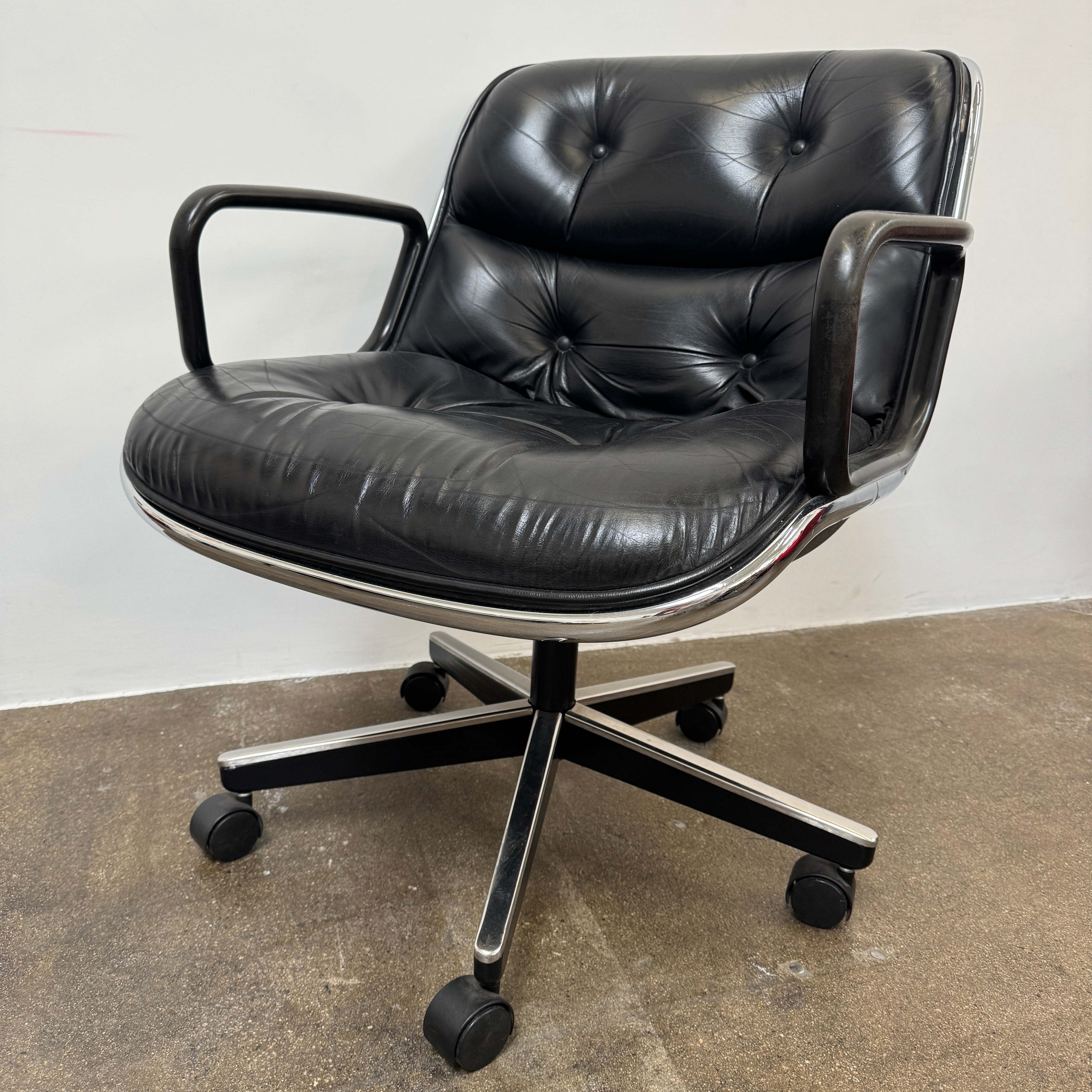 A DWR Pollock Executive Chair with a chrome frame and five wheels is shown from the back, set on a concrete floor against a plain white wall.