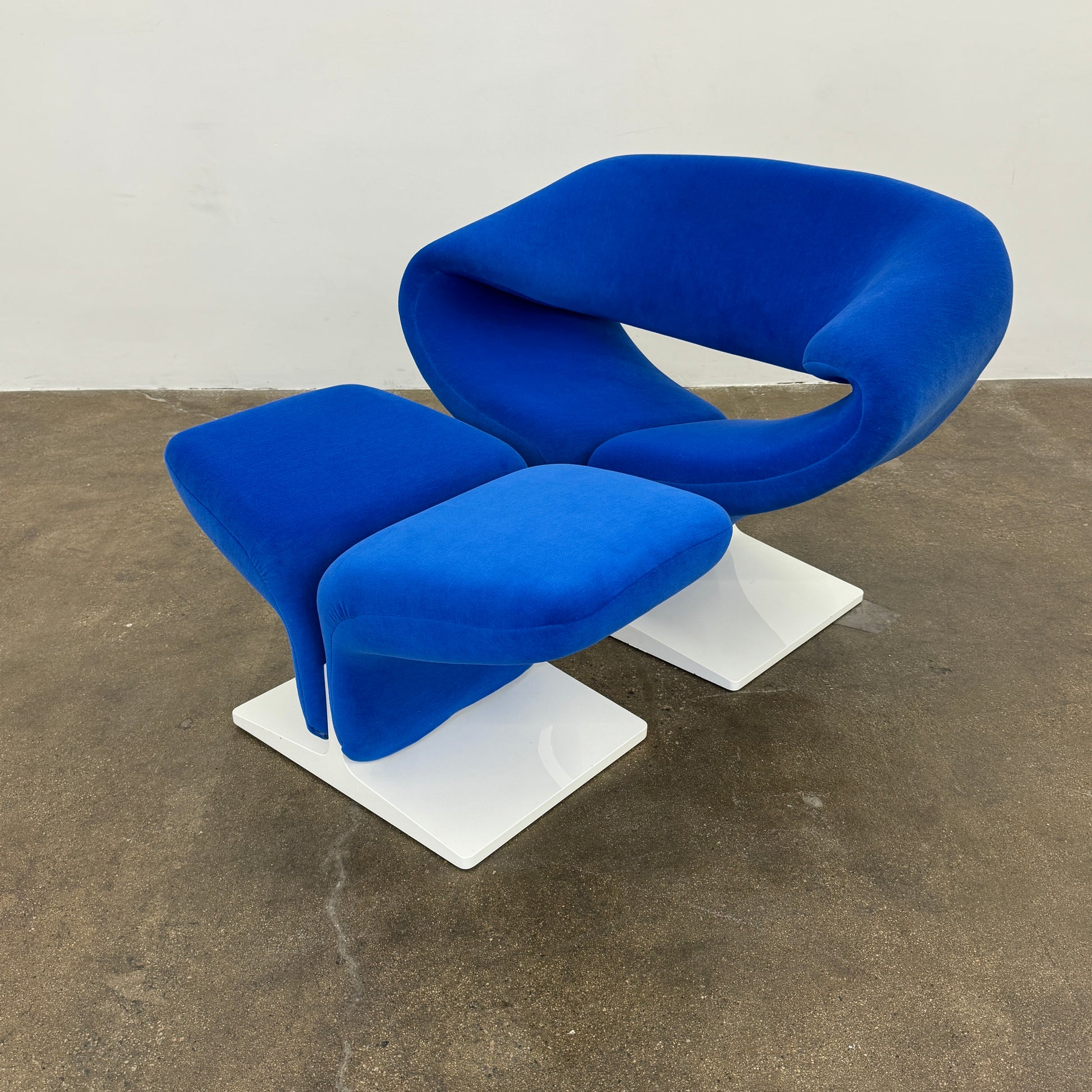 The Artifort Ribbon Lounge Chair & Ottoman by Artifort features a modern blue design with a sculptural curved backrest and split seat on a white base, seen here against a plain white background.