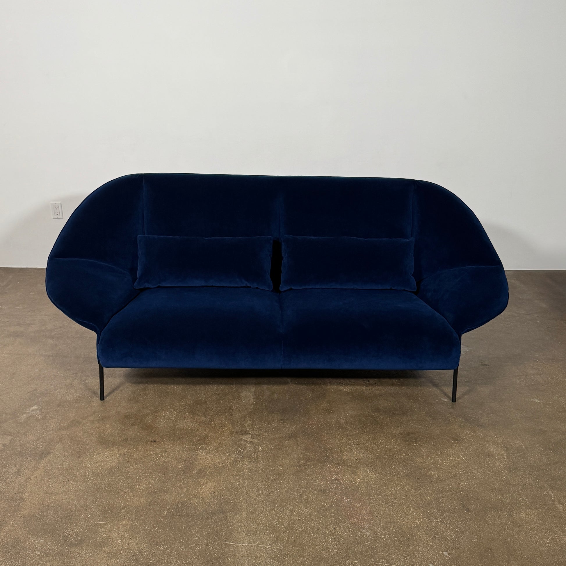The Ligne Roset Paipaï Sofa by Ligne Roset features deep blue velvet, curved edges, slim black legs, premium foam construction, and two matching rectangular cushions for a cocoon-like seat against a light background.