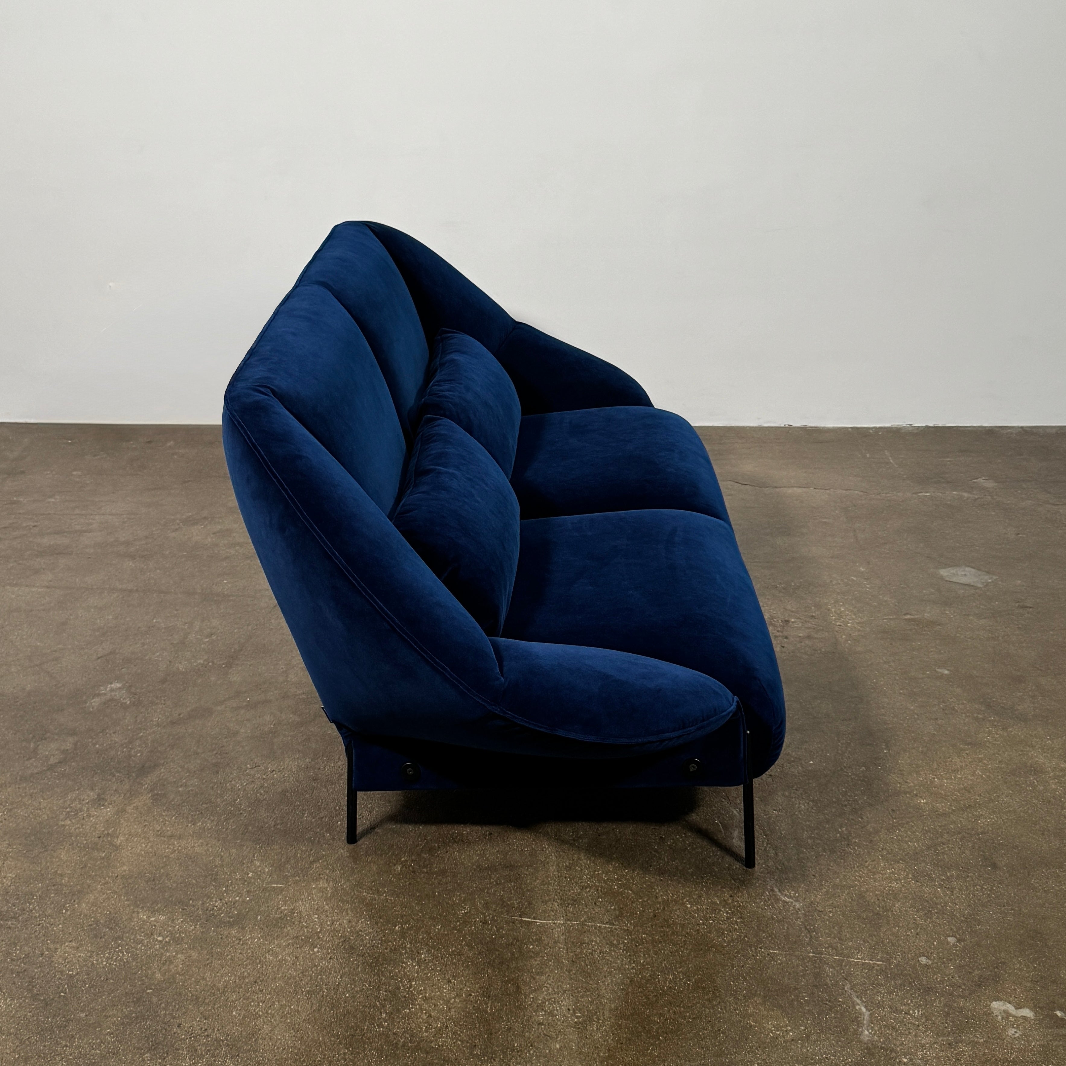 The Ligne Roset Paipaï Sofa by Ligne Roset features deep blue velvet, curved edges, slim black legs, premium foam construction, and two matching rectangular cushions for a cocoon-like seat against a light background.