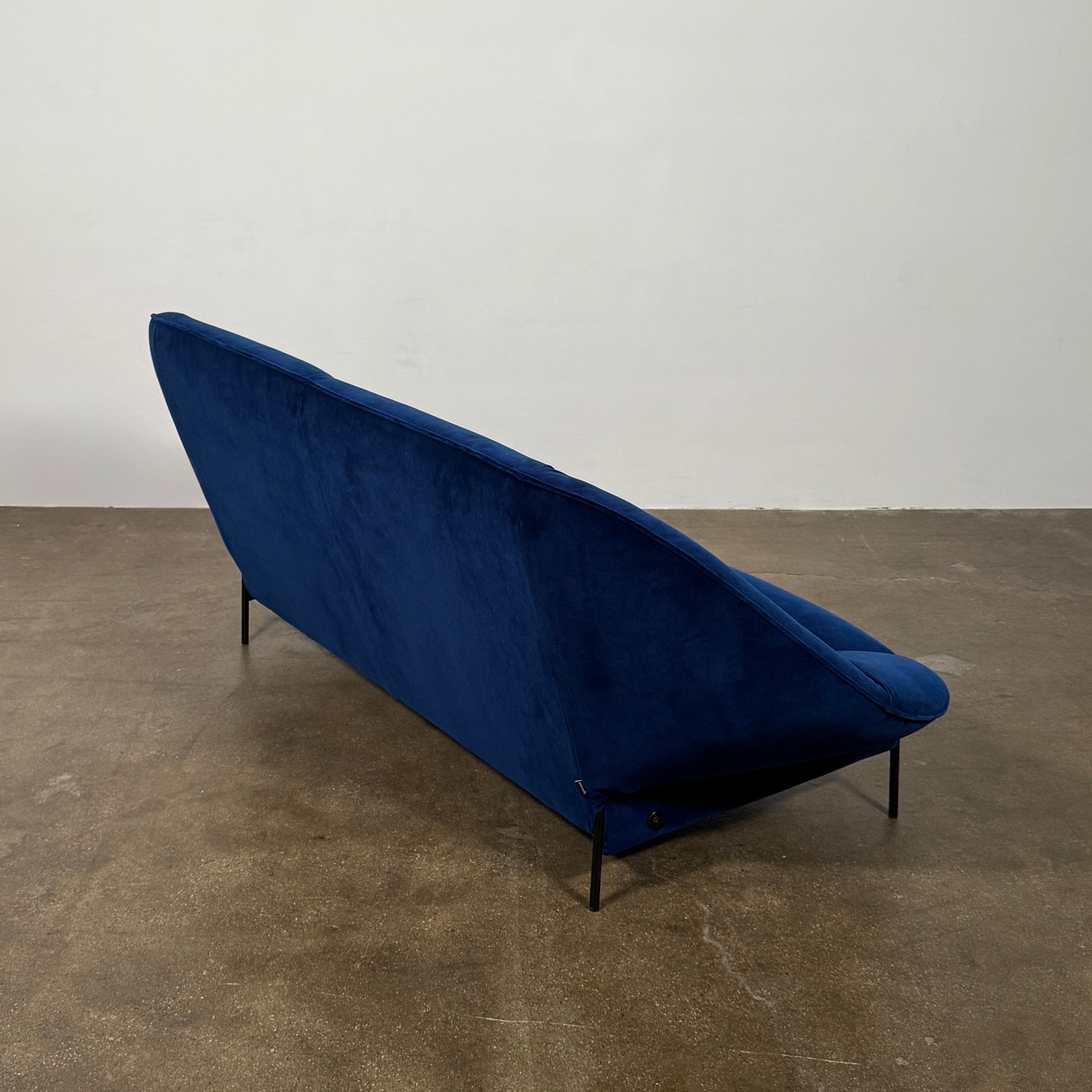 The Ligne Roset Paipaï Sofa by Ligne Roset features deep blue velvet, curved edges, slim black legs, premium foam construction, and two matching rectangular cushions for a cocoon-like seat against a light background.