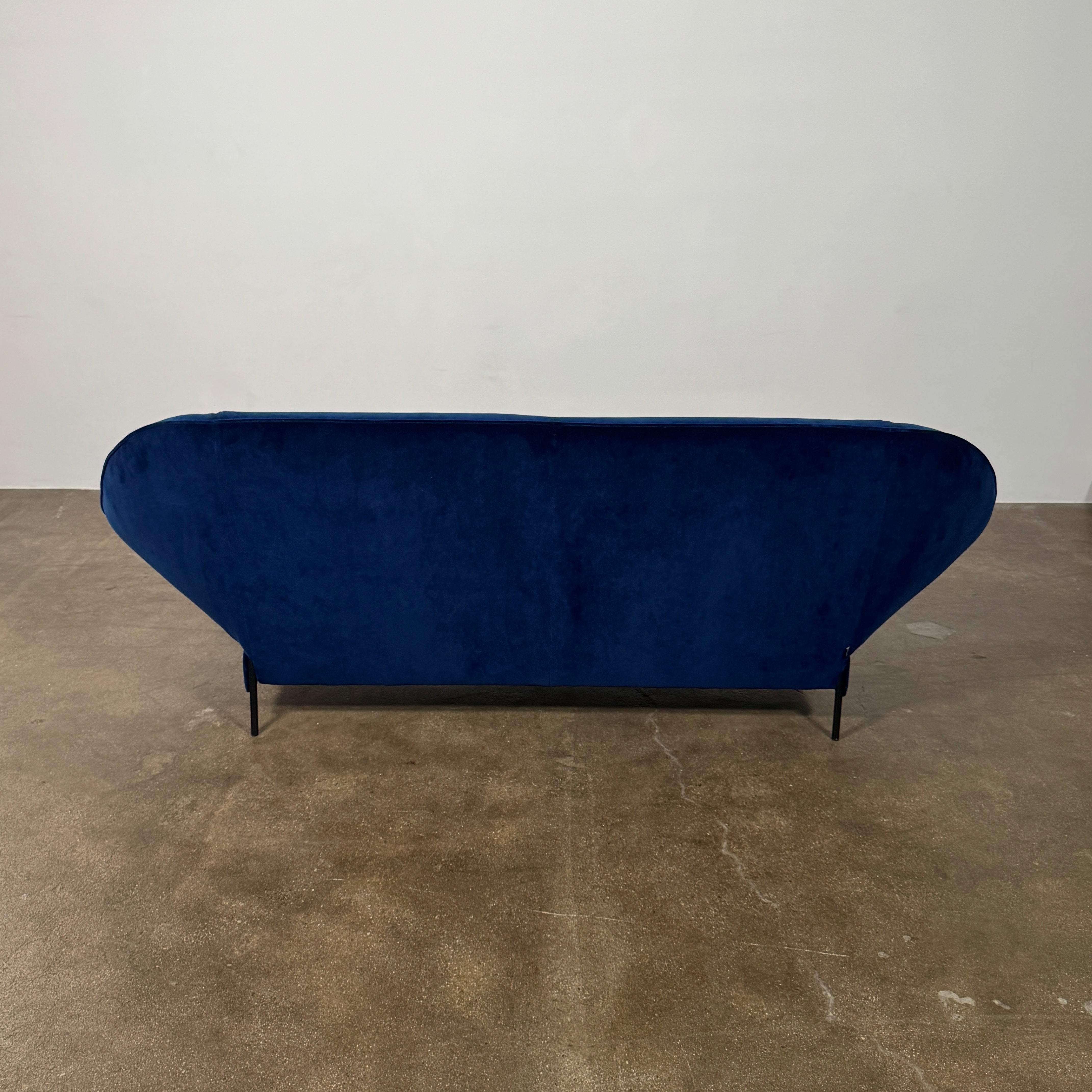 The Ligne Roset Paipaï Sofa by Ligne Roset features deep blue velvet, curved edges, slim black legs, premium foam construction, and two matching rectangular cushions for a cocoon-like seat against a light background.